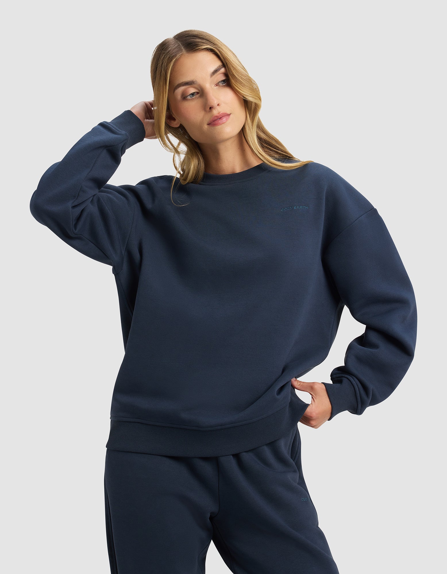 A woman with long blonde hair models the Cozy Earth Women's Cityscape 2.0 Crewneck in navy, paired with matching sweatpants. She stands with one hand behind her head and the other on her hip, gazing off to the side.|Color:Eclipse