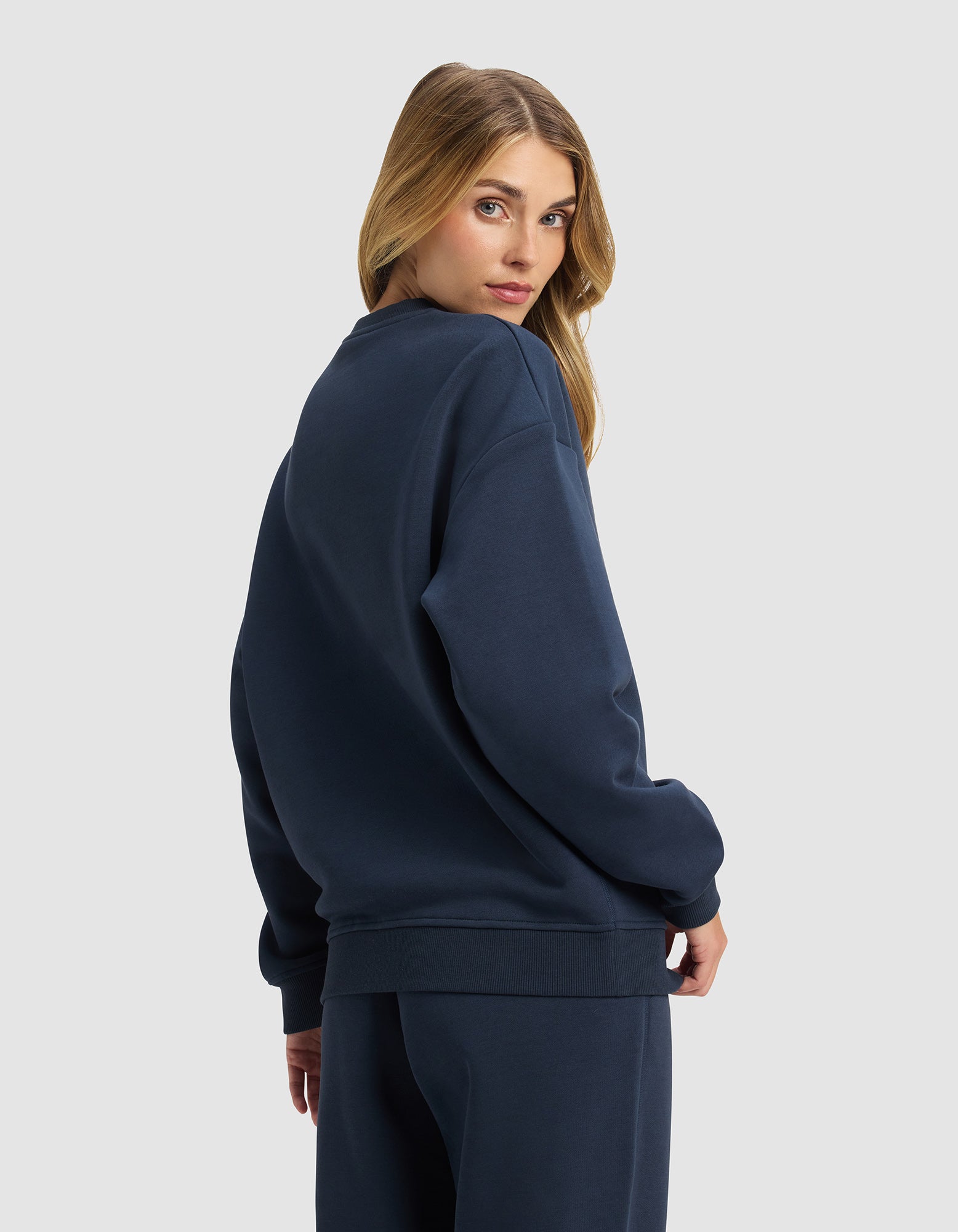 A woman with long blonde hair models the Cozy Earth Women's Cityscape 2.0 Crewneck in navy blue, paired with matching pants, as she stands against a light gray background and glances over her shoulder at the camera.