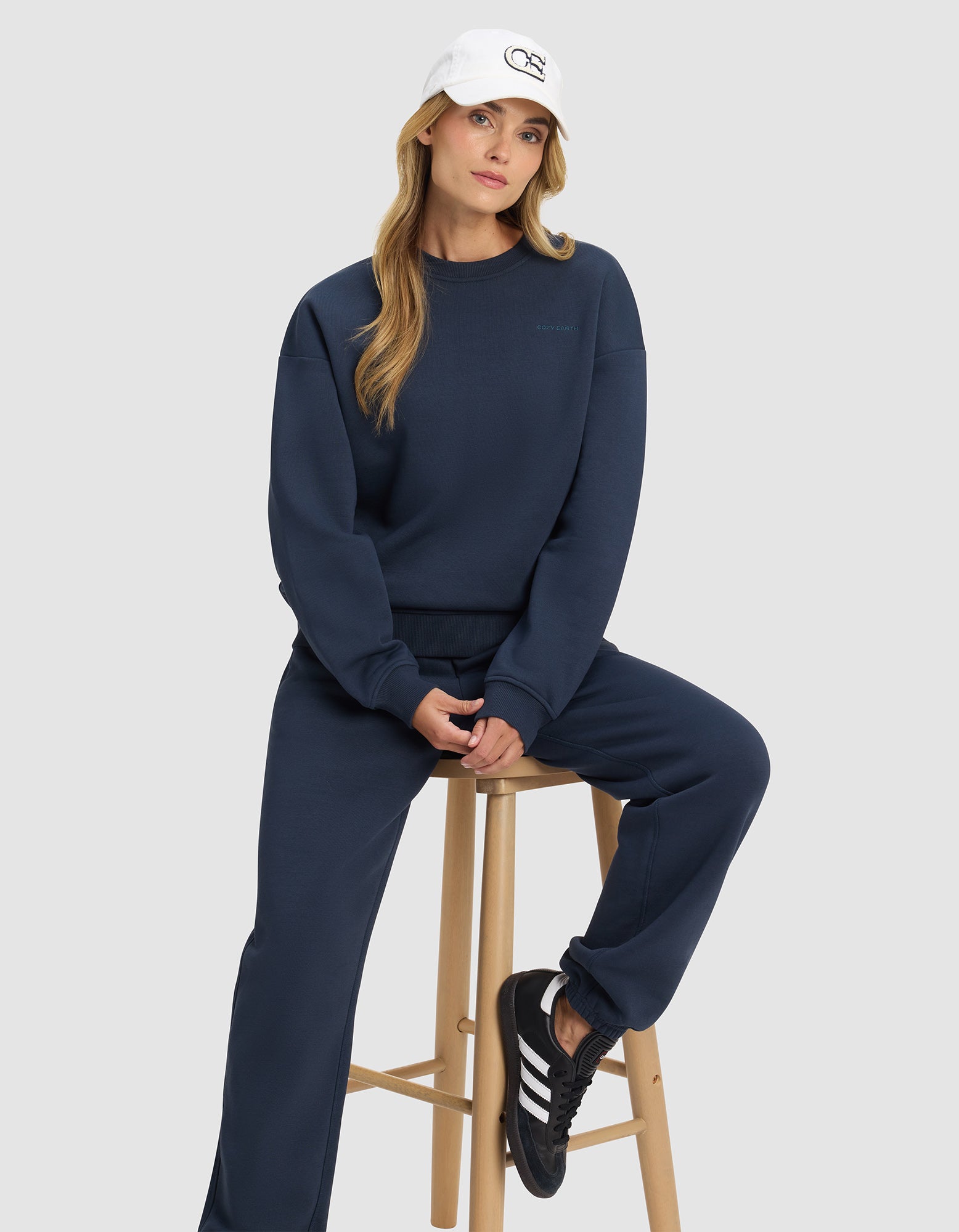 A woman in the Cozy Earth Women's Cityscape 2.0 Crewneck and matching navy sweatpants sits on a wooden stool. She wears a white cap, has long blonde hair, and sports black Adidas sneakers with white stripes against a light gray background.
