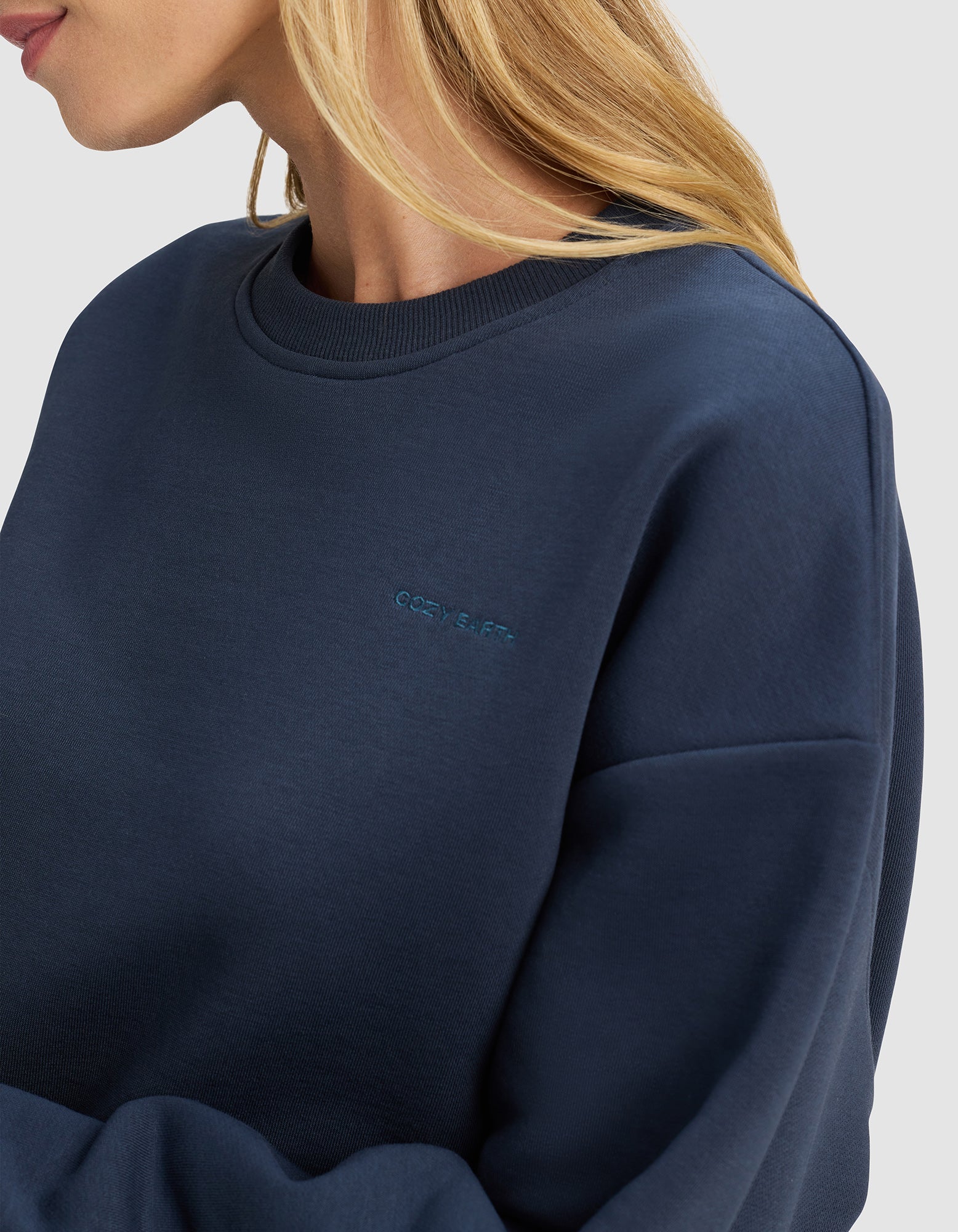 A woman with blonde hair models the Cozy Earth Women's Cityscape 2.0 Crewneck, a dark blue sweatshirt with subtle chest embroidery, standing against a plain light background.