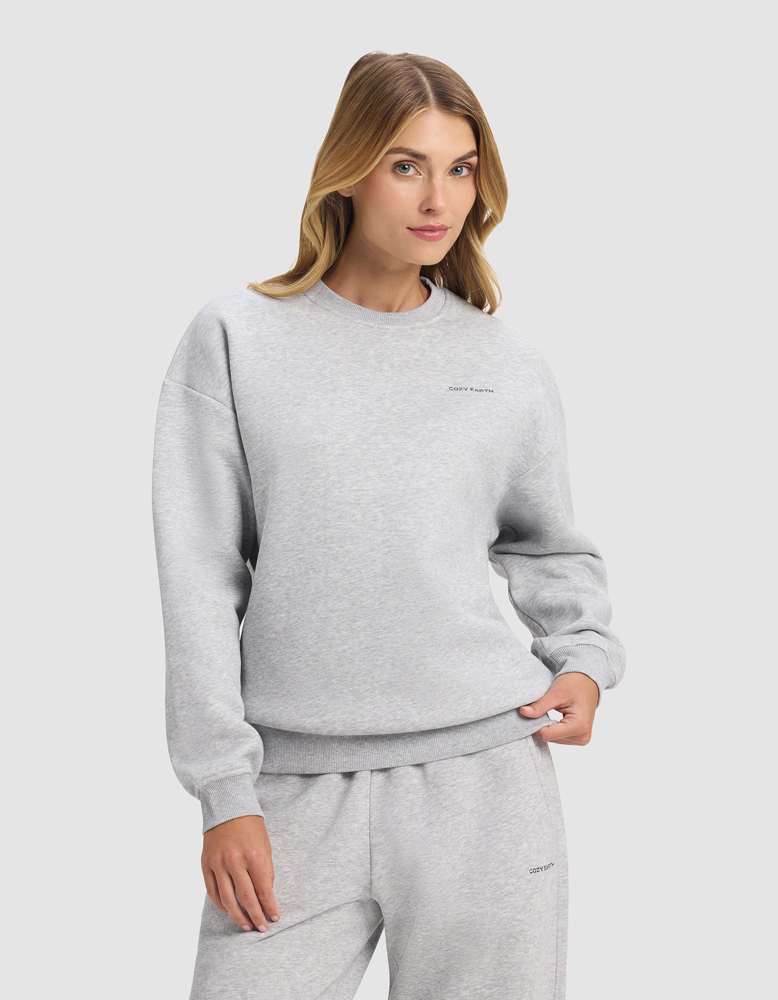 A woman with long blonde hair models the Cozy Earth Women's Cityscape 2.0 Crewneck in light gray, paired with matching sweatpants, standing with one hand in her pocket against a plain light background.