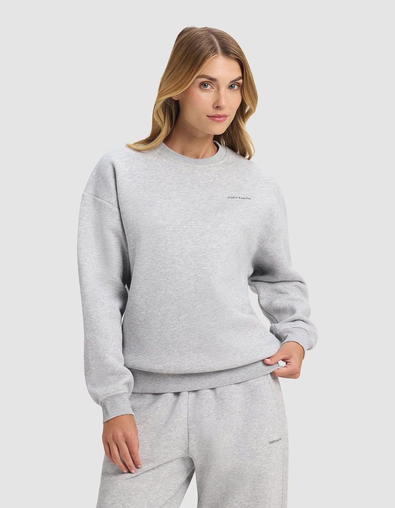 A woman with long blonde hair models the Cozy Earth Women's Cityscape 2.0 Crewneck in light gray, paired with matching sweatpants, standing with one hand in her pocket against a plain light background.