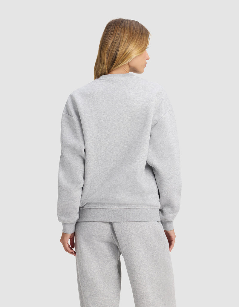 A woman with blonde hair faces away, wearing the Cozy Earth Women's Cityscape 2.0 Crewneck in light gray with matching sweatpants against a plain light background.