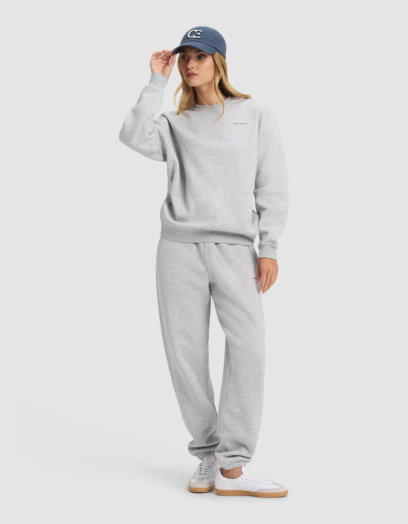 A woman in Cozy Earth's Women's Cityscape 2.0 Crewneck poses against a plain background, paired with matching sweatpants, white sneakers, and a navy blue cap as she touches the brim and gazes to the side.