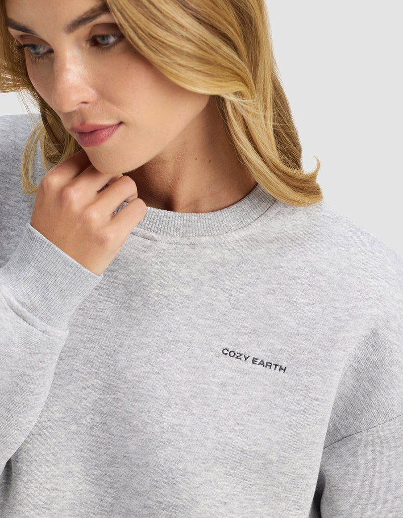 A woman with wavy blonde hair wears the Cozy Earth Women's Cityscape 2.0 Crewneck in light gray, featuring the brand embroidered on the chest, as she gazes down thoughtfully and touches her chin.