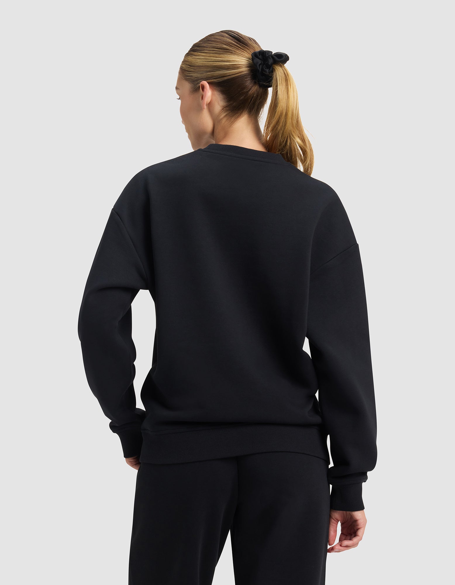 A woman with blonde hair in a ponytail, secured with a black scrunchie, wears the Cozy Earth Women's Cityscape 2.0 Crewneck and black pants. She stands facing away from the camera against a plain light gray background.