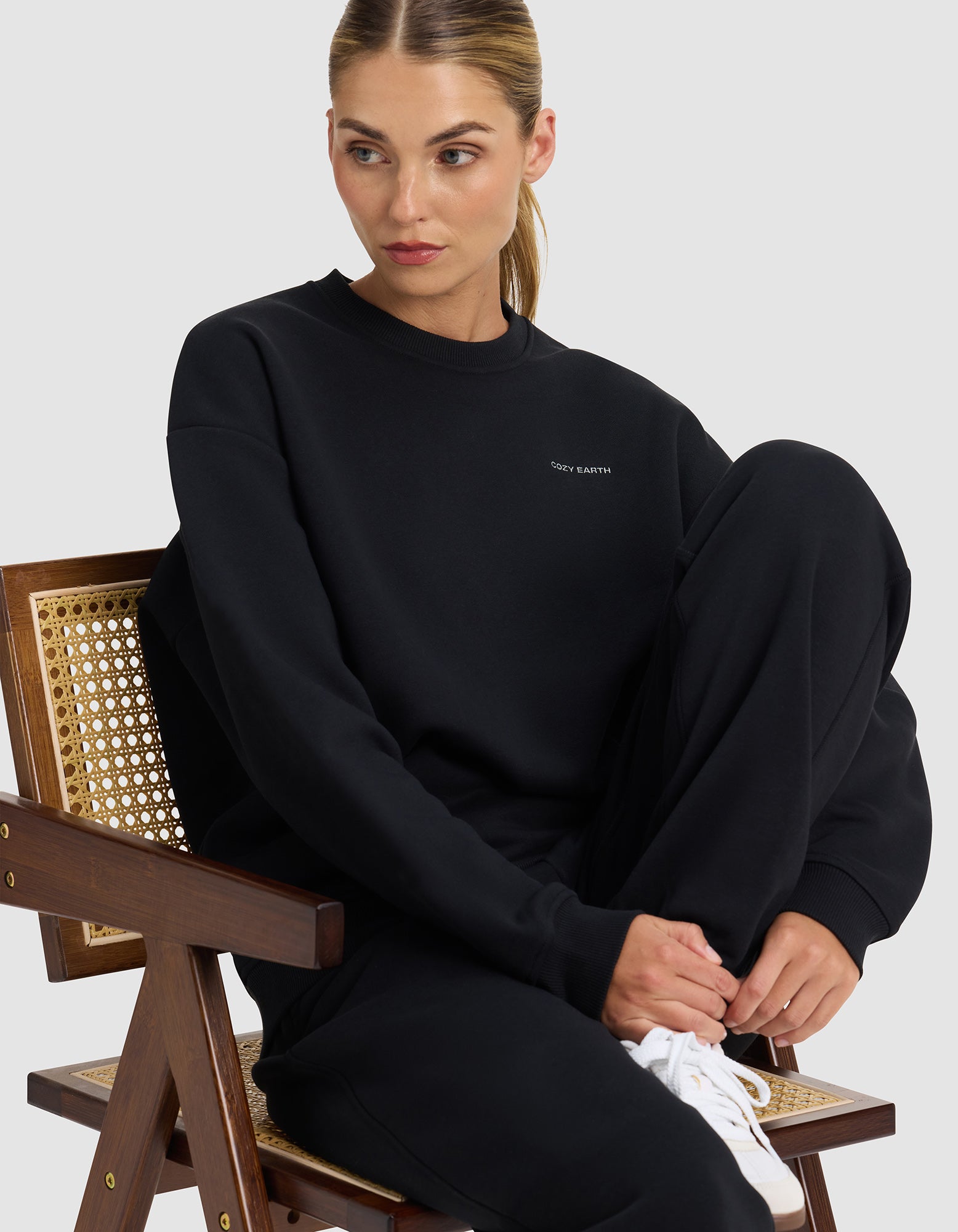 A woman with light brown hair tied back sits sideways on a wooden chair, wearing Cozy Earth's Women's Cityscape 2.0 Crewneck in black with matching sweatpants and white sneakers, looking to her left.