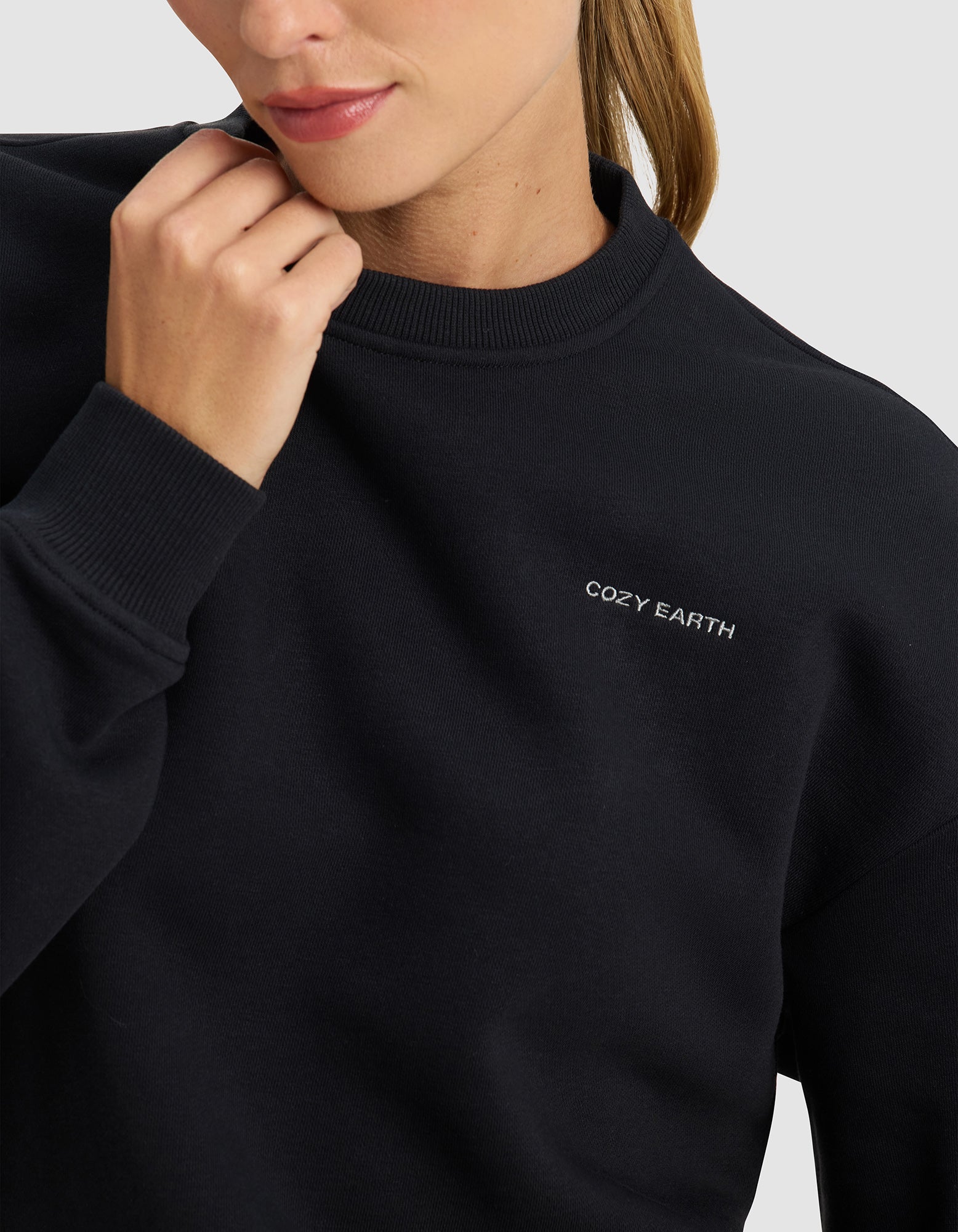 A woman wears the Cozy Earth Women's Cityscape 2.0 Crewneck in black, featuring "COZY EARTH" embroidered in white on the chest, shown from the shoulders up with her hand touching her chin.