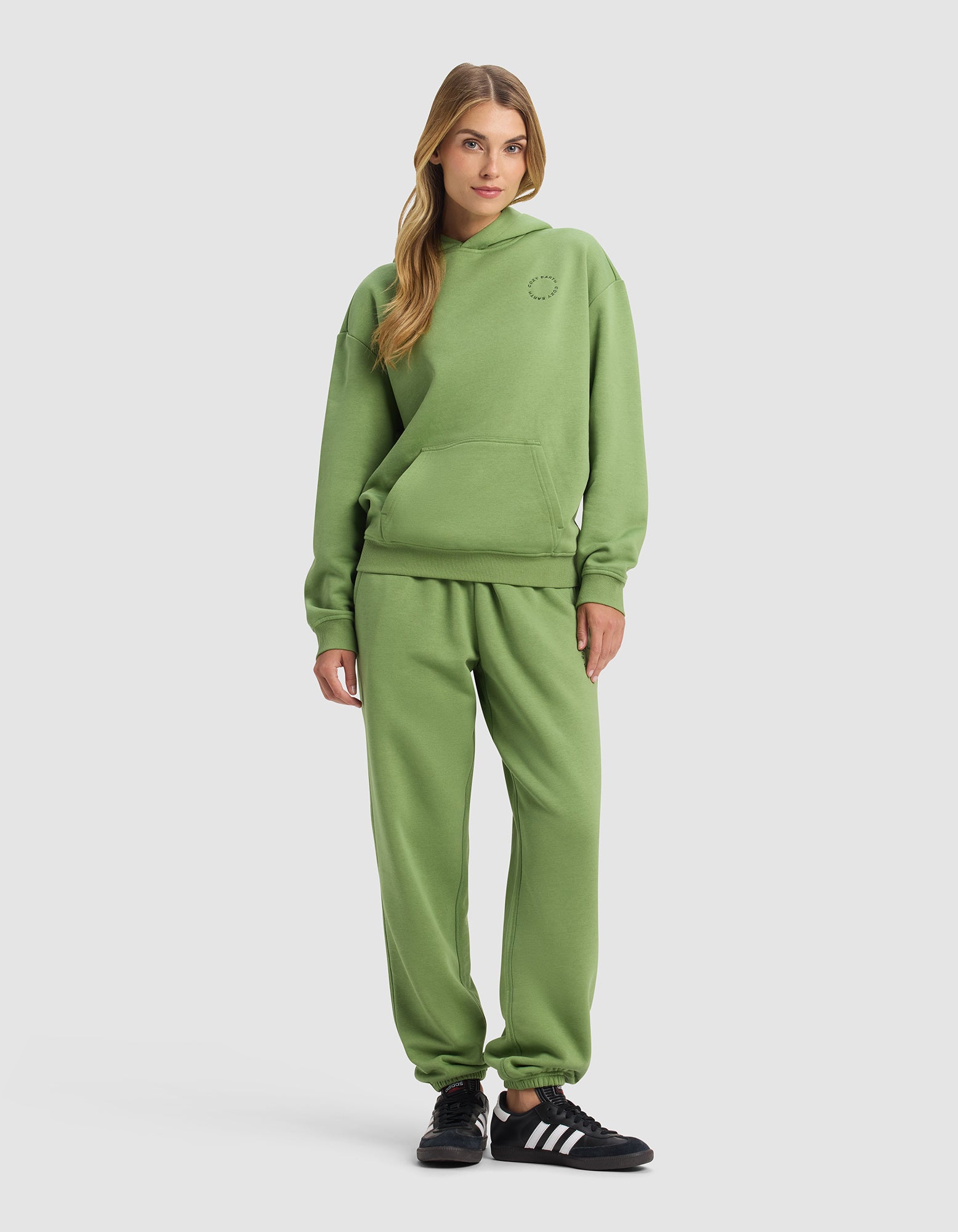 A woman wears the Cozy Earth Women's Cityscape 2.0 Hoodie & Sweatpant Set in green with black Adidas sneakers, standing with hands relaxed at her sides against a plain light gray background.