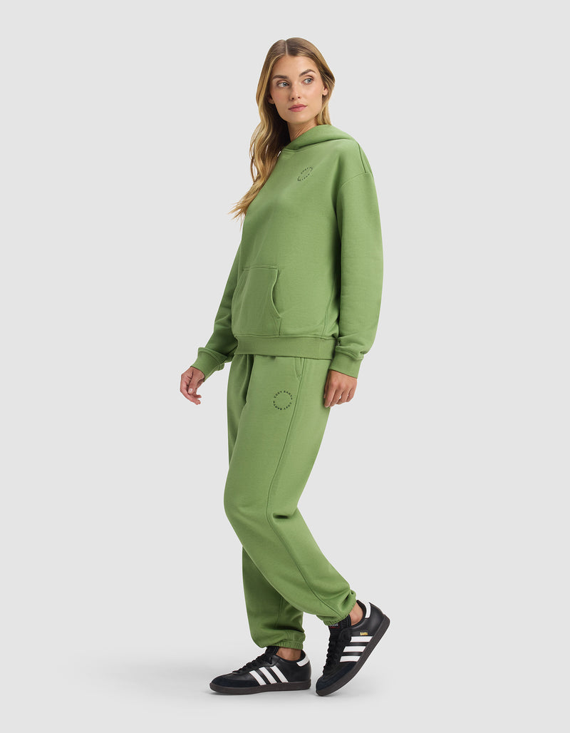 A woman stands against a plain light background wearing the Cozy Earth Women's Cityscape 2.0 Hoodie & Sweatpant Set in light green with small circular designs, paired with black Adidas sneakers.