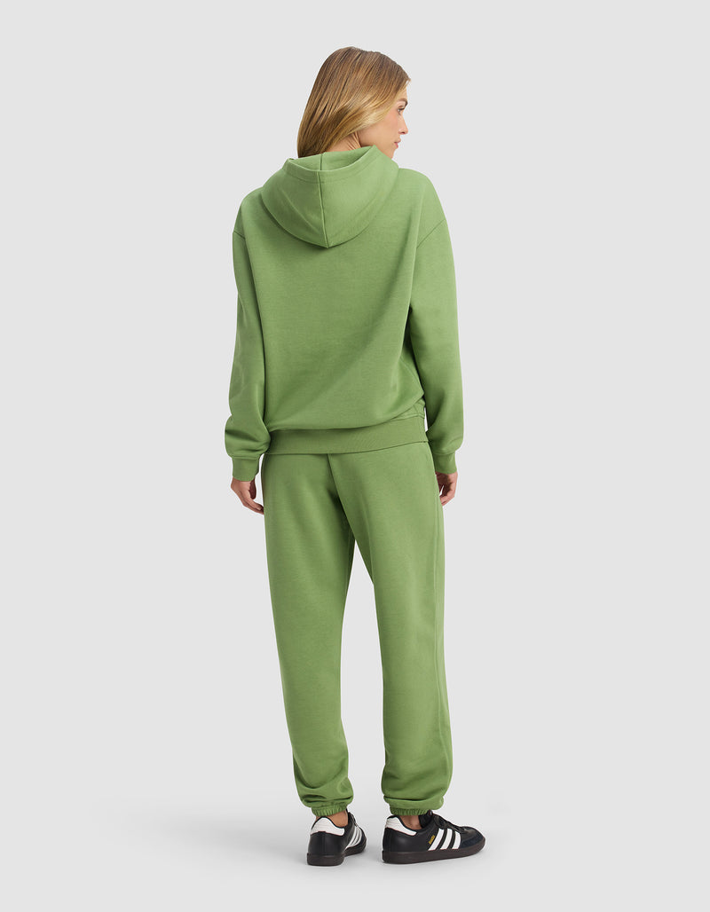 A person with blonde hair stands facing away, wearing the Cozy Earth Women's Cityscape 2.0 Hoodie & Sweatpant Set in green and black and white sneakers, set against a plain light background.
