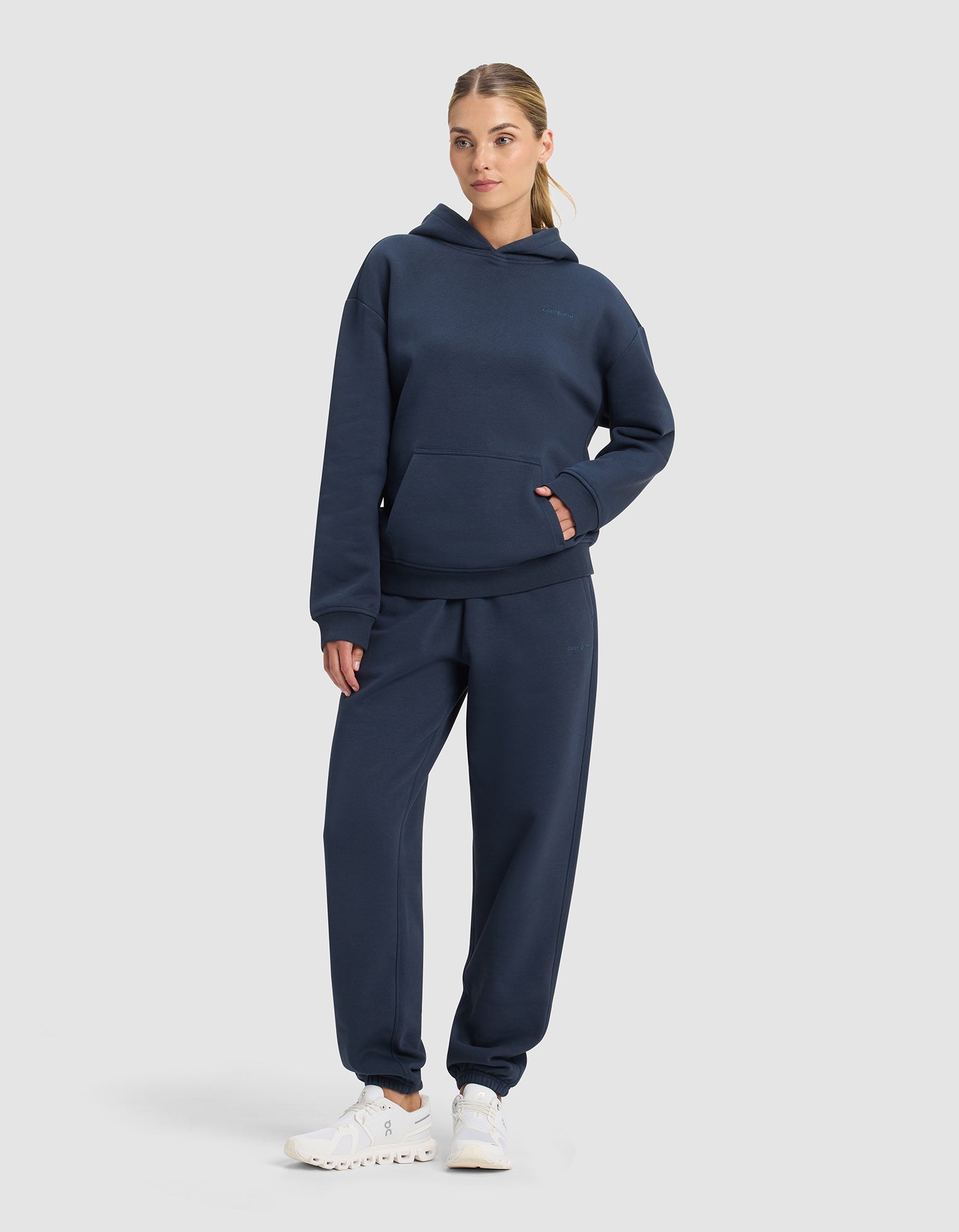 A woman poses against a plain backdrop in the Cozy Earth Women's Cityscape 2.0 Hoodie & Sweatpant Set, paired with white sneakers. She has one hand in her pocket and the other on her hip.|Color:Eclipse