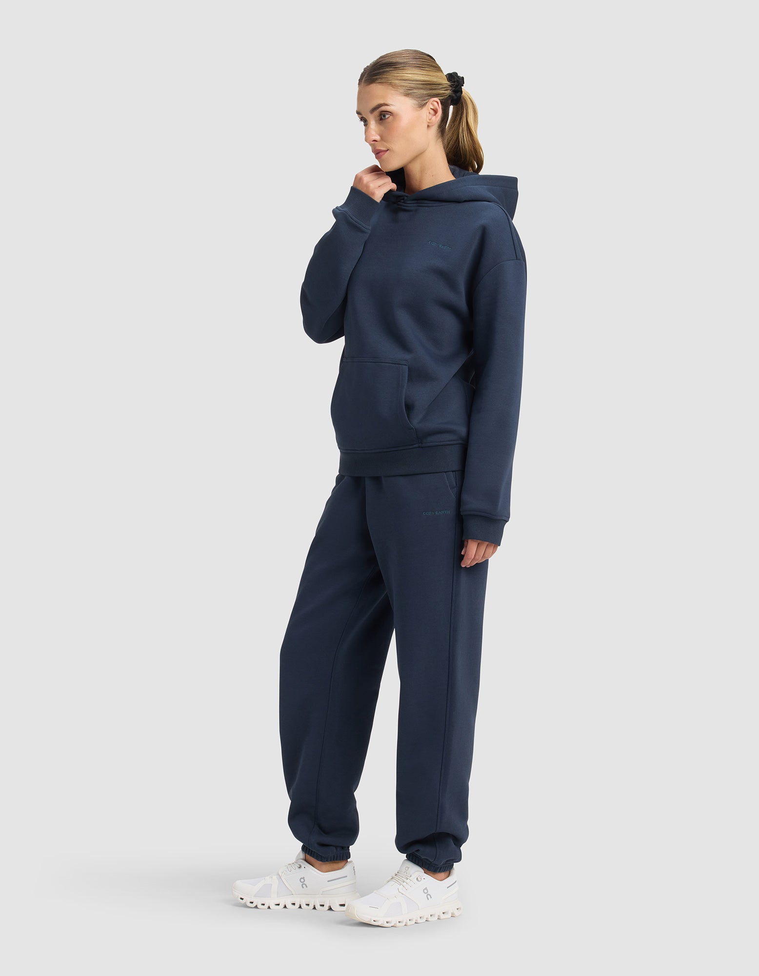 A woman poses against a plain background wearing the Cozy Earth Women's Cityscape 2.0 Hoodie & Sweatpant Set in navy blue with white sneakers. Her hair is tied back in a ponytail, and she holds her hand near her chin.|Color:Eclipse
