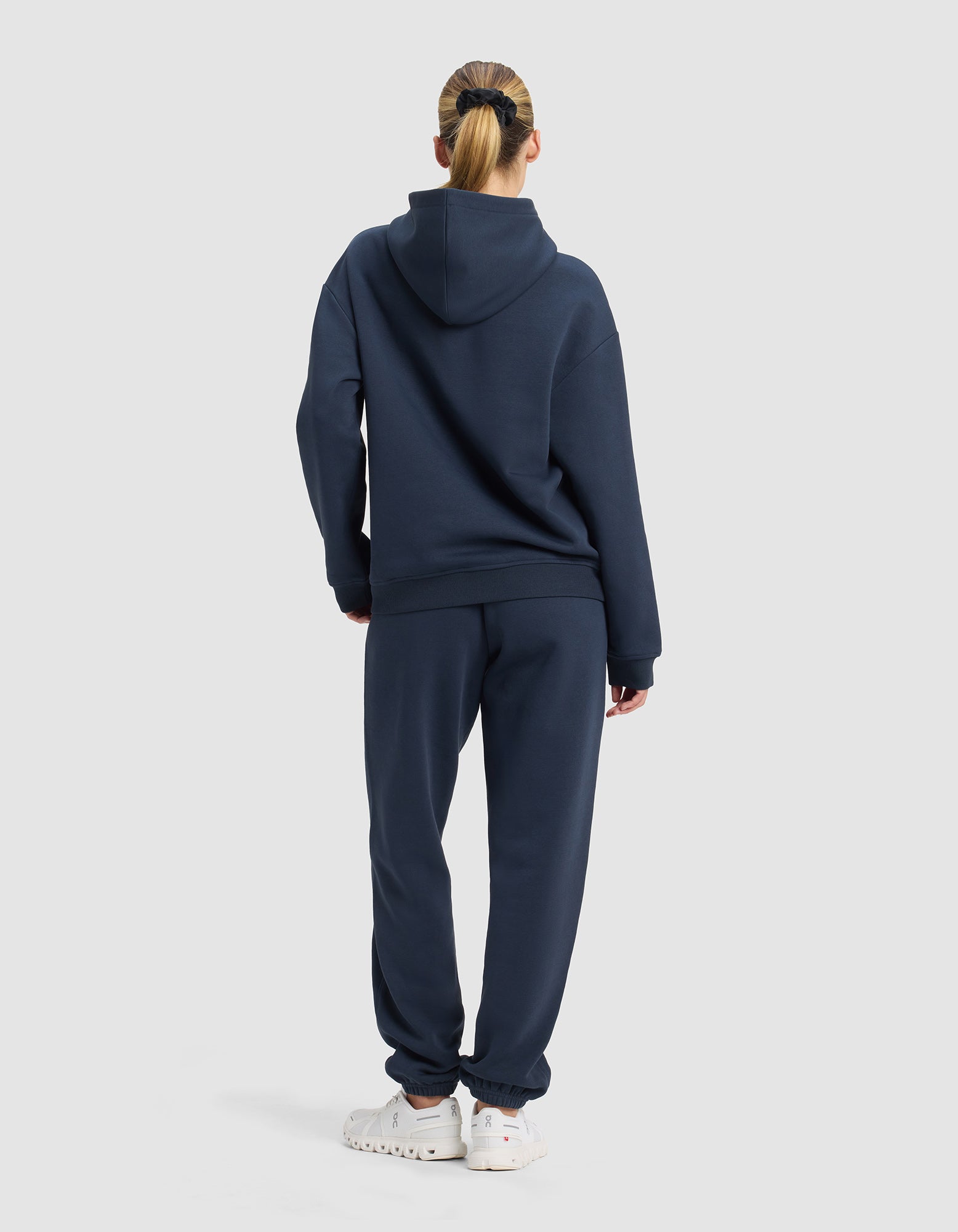 A woman with blonde hair in a ponytail stands facing away, wearing the Cozy Earth Women's Cityscape 2.0 Hoodie & Sweatpant Set in navy blue with white sneakers, set against a plain light gray background.|Color:Eclipse