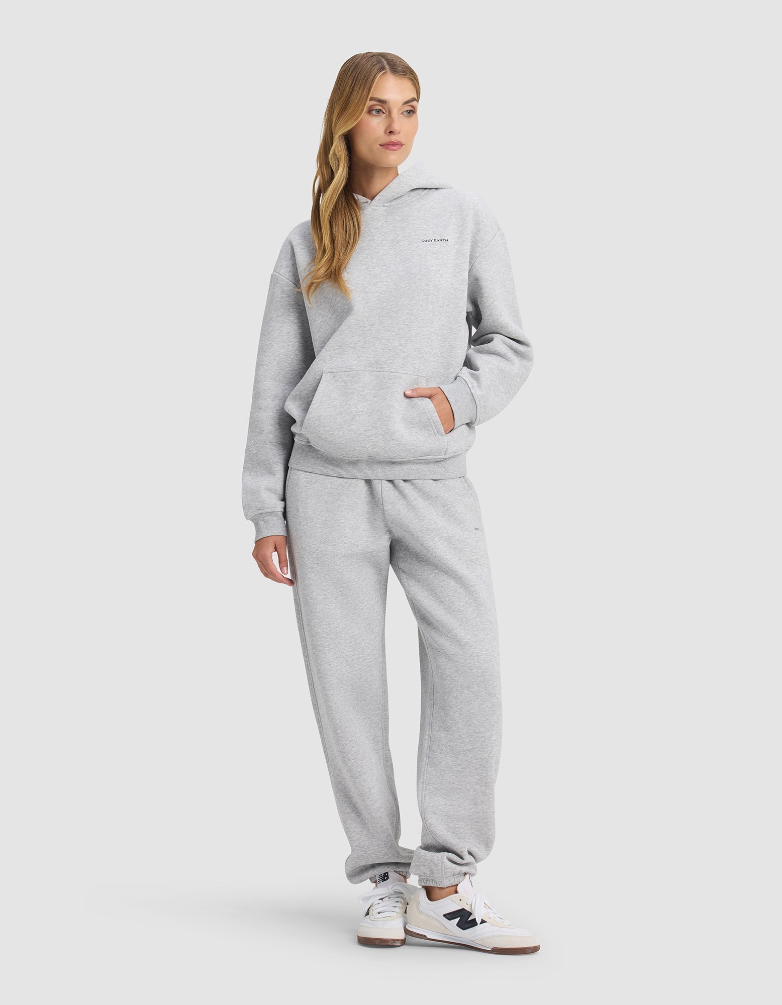 A woman wears the Cozy Earth Women's Cityscape 2.0 Hoodie & Sweatpant Set in light gray, paired with white sneakers with black accents. She stands with one hand in her pocket against a plain light background, her long hair loose.