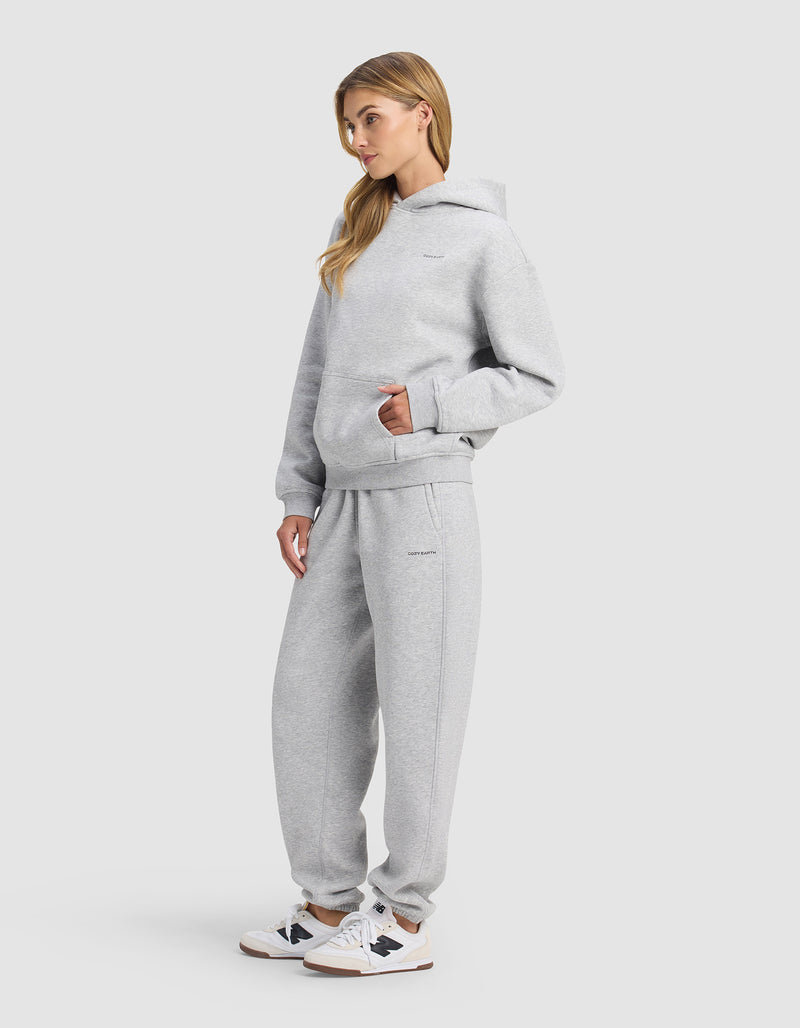 A woman with long blonde hair stands in a relaxed pose, hands in the front pocket, wearing Cozy Earth's Women's Cityscape 2.0 Hoodie & Sweatpant Set in light gray with white sneakers against a plain light gray background.