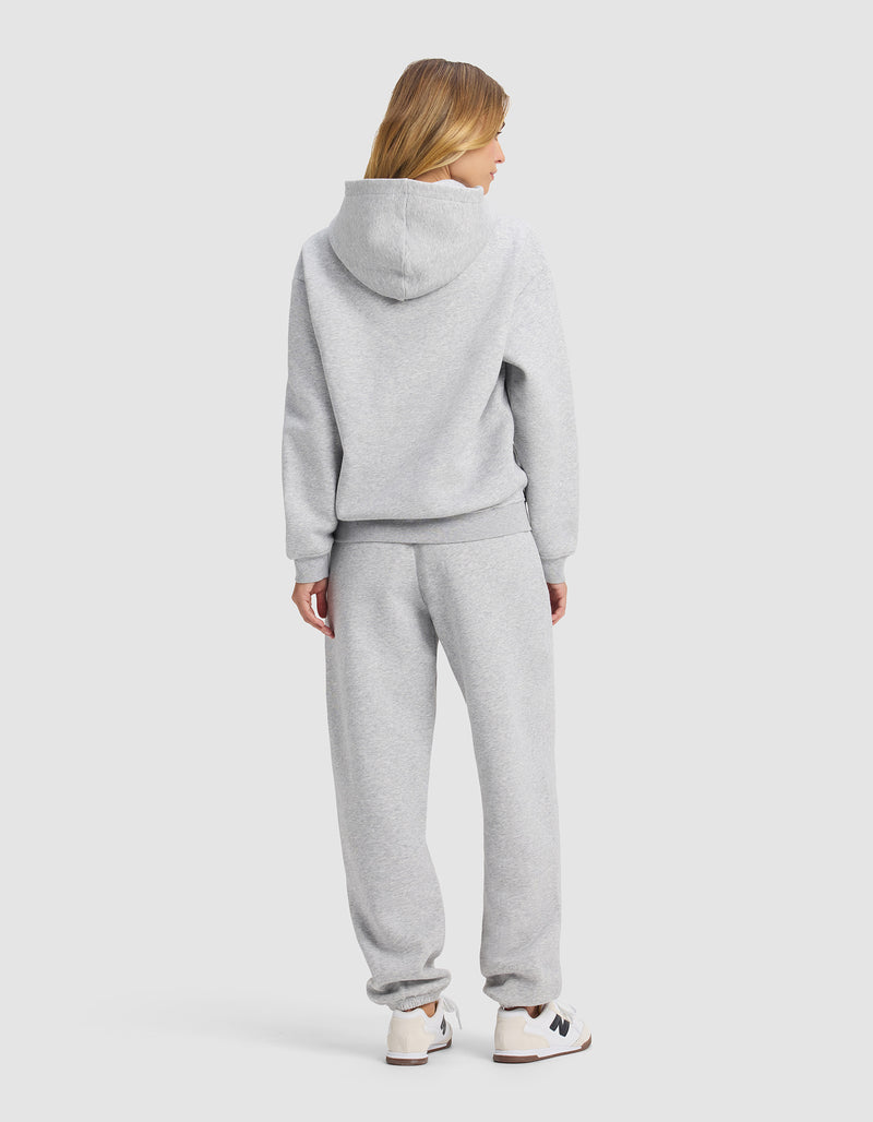 A woman with blonde hair, seen from behind, wears the Cozy Earth Women's Cityscape 2.0 Hoodie & Sweatpant Set in light gray with white sneakers against a plain light gray background.