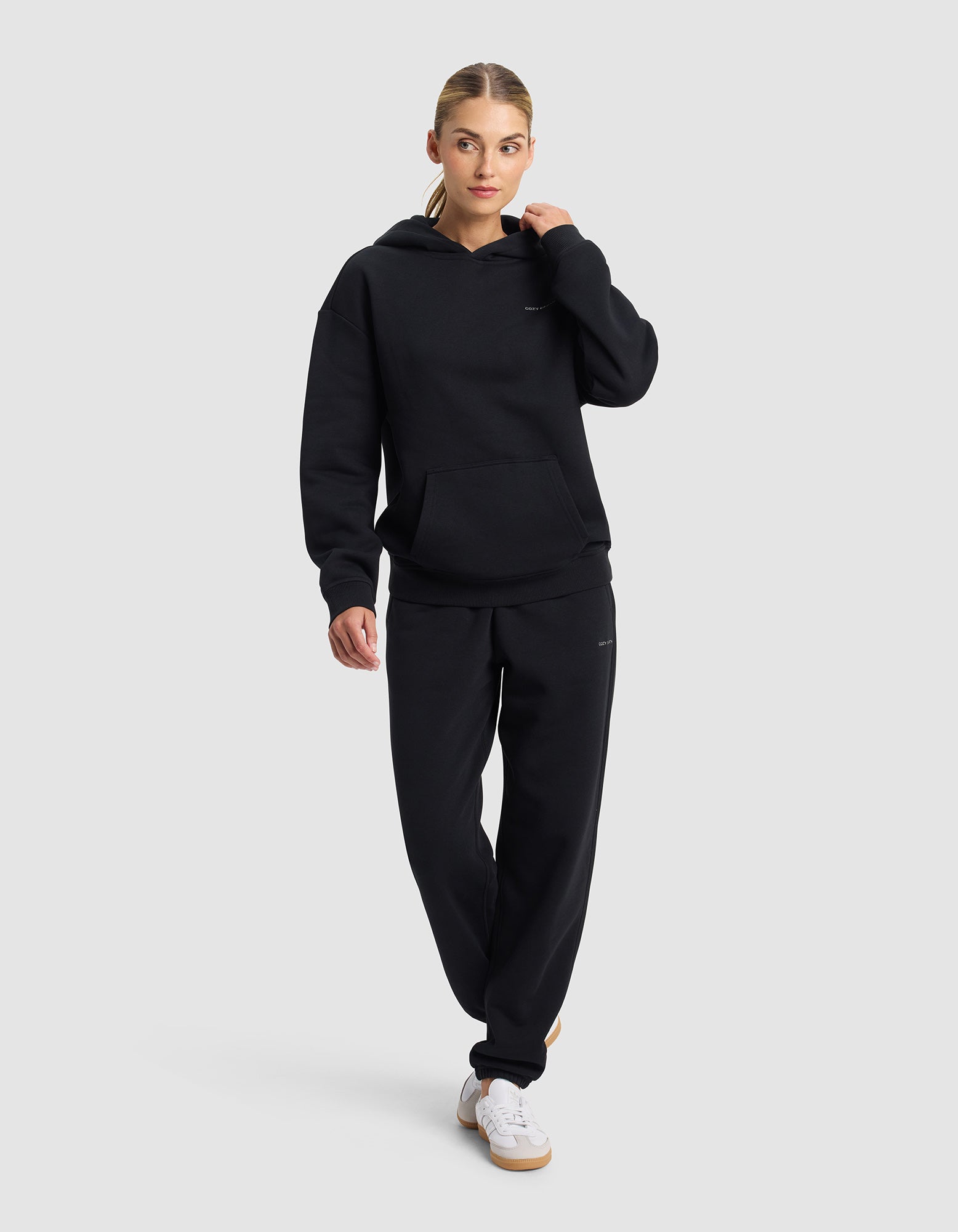 A woman in the Cozy Earth Women's Cityscape 2.0 Hoodie & Sweatpant Set stands against a light gray background, wearing black with white sneakers, her hair tied back and one hand touching her hood.|Color:Jet Black