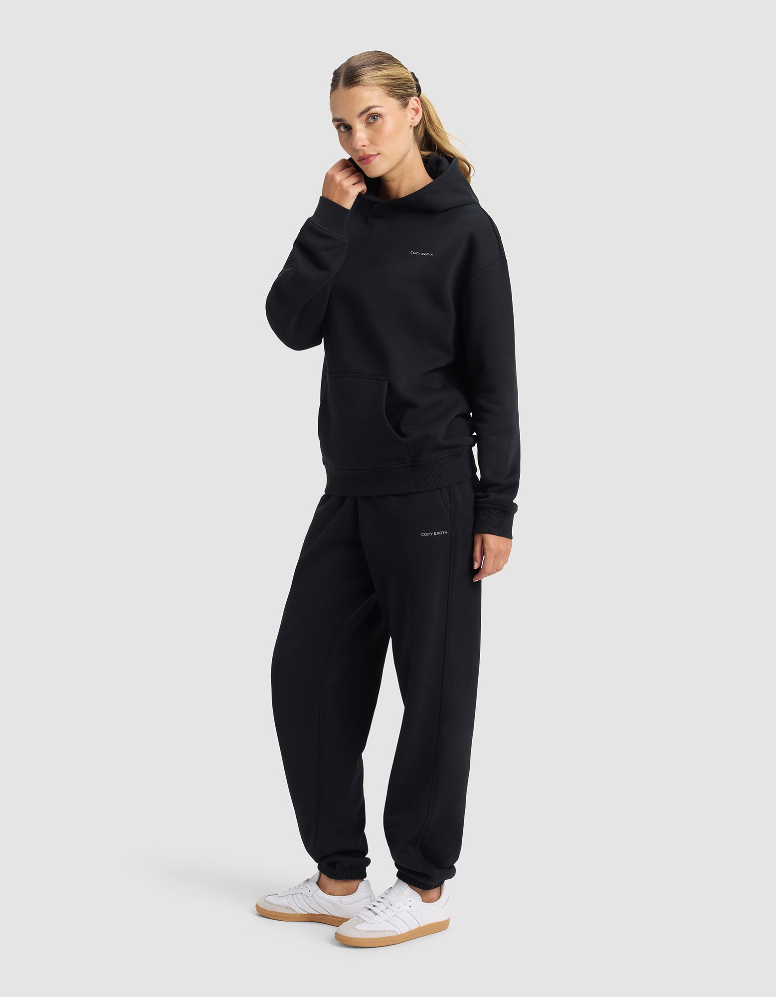 A woman models the Cozy Earth Women's Cityscape 2.0 Hoodie & Sweatpant Set in black with small white text, paired with white sneakers, against a plain background. Her hair is tied back as she looks at the camera.