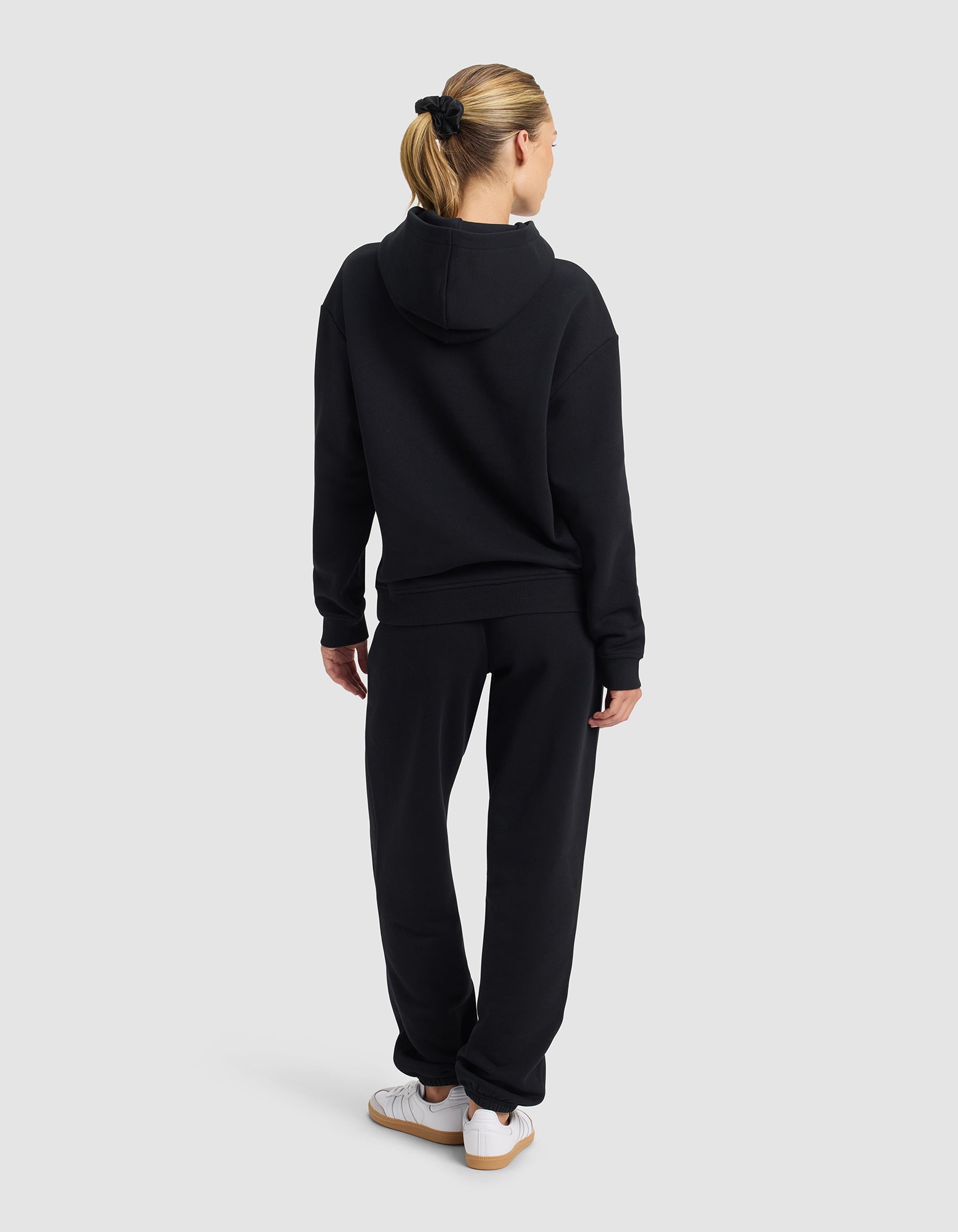 A person with blonde hair in a ponytail, wearing Cozy Earth's Women's Cityscape 2.0 Hoodie & Sweatpant Set in black and white sneakers, stands facing away against a plain light gray background.