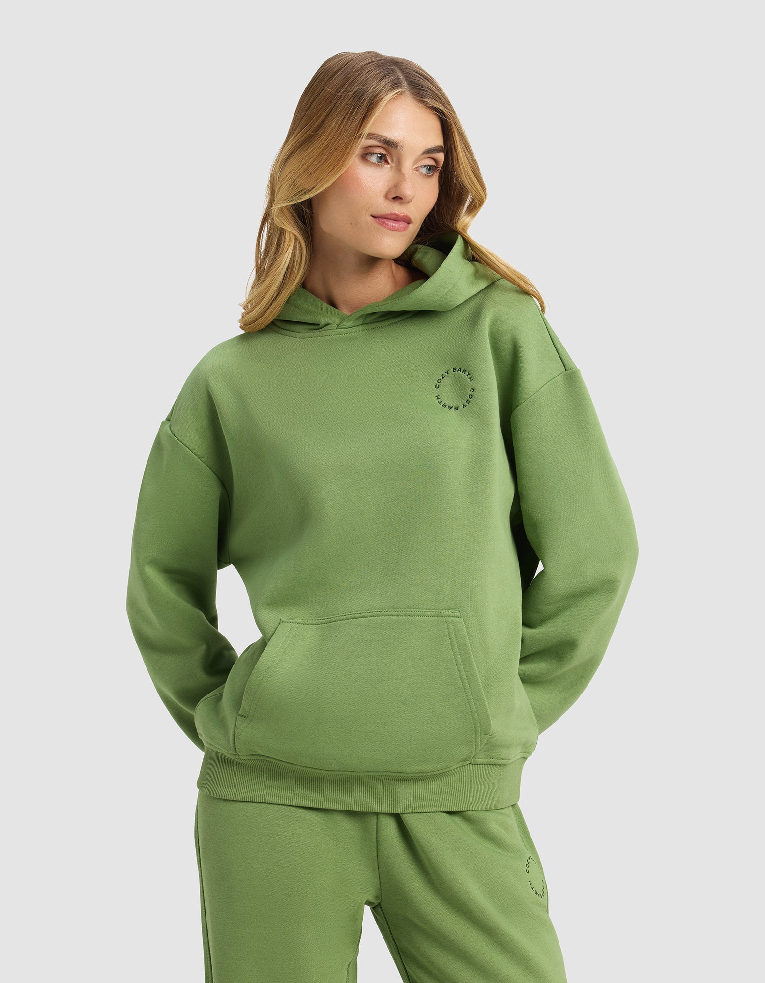 A woman with long blonde hair models the Cozy Earth Women's Cityscape 2.0 Hoodie in green, paired with matching sweatpants, standing with hands in her hoodie pocket against a plain light gray background.|Color:Clover