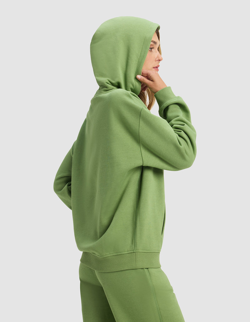 A woman in profile wears the Cozy Earth Women's Cityscape 2.0 Hoodie and matching sweatpants in green, standing against a plain light background with her hood up and one hand touching her chin.