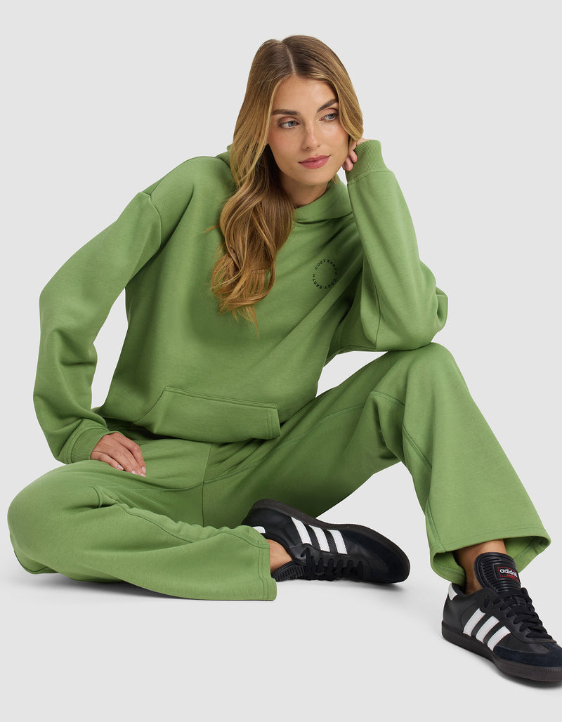 A woman with long, wavy hair sits on the floor wearing Cozy Earth’s Women’s Cityscape 2.0 Hoodie in green and matching sweatpants, paired with black Adidas sneakers. She rests her head on one hand and looks to the side.