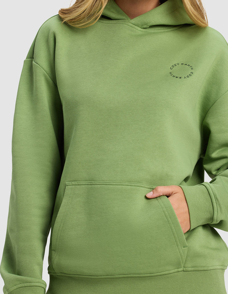 A woman wears the Cozy Earth Women's Cityscape 2.0 Hoodie in light green, featuring a front pocket and small circular "COZY EARTH" text on the chest. Only her lower face, neck, and upper body are visible.