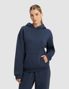 A woman with blonde hair in a low ponytail wears the Cozy Earth Women's Cityscape 2.0 Hoodie in navy blue, standing against a light gray background with one hand in her hoodie pocket.|Color:Eclipse