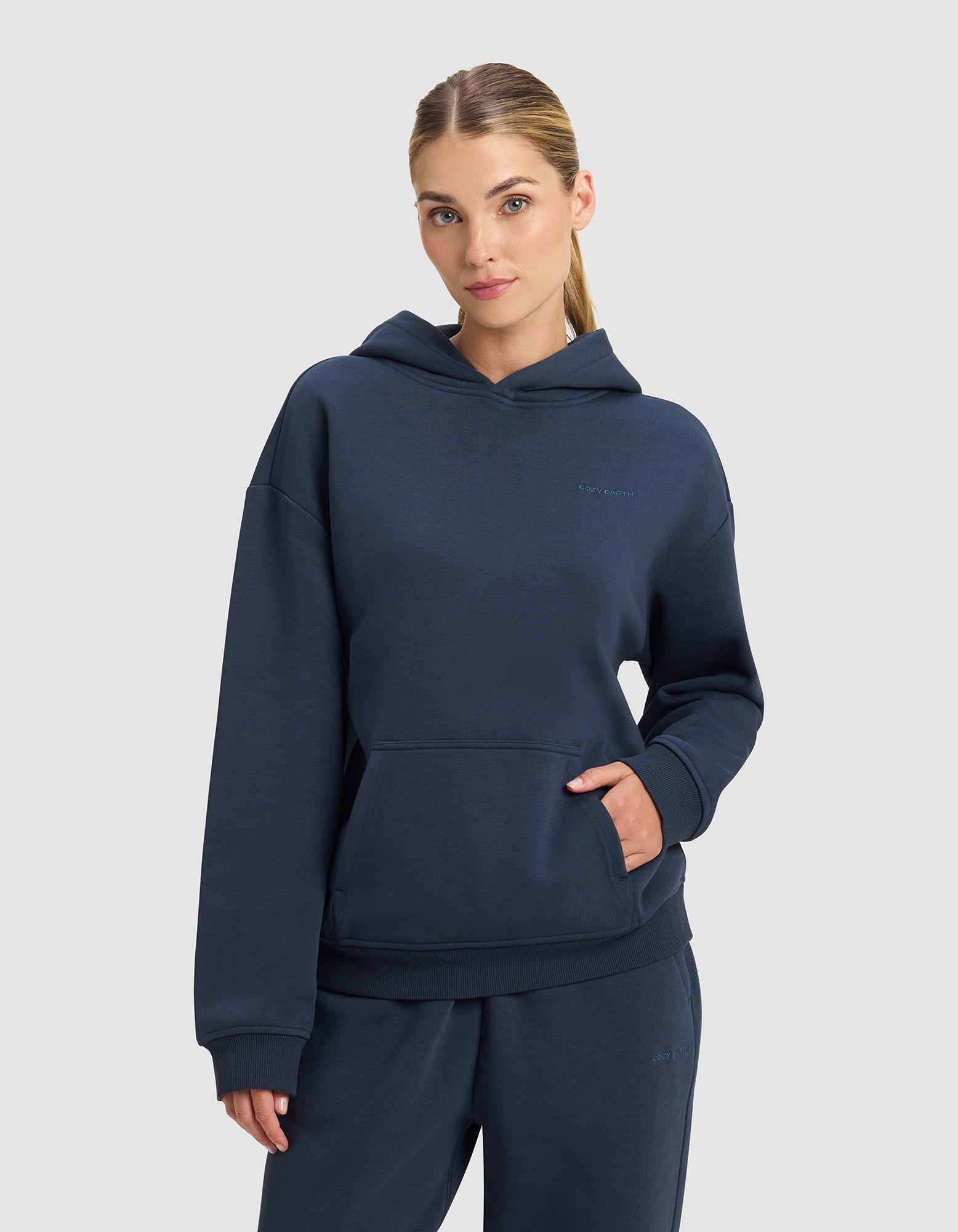 A woman with blonde hair in a low ponytail wears the Cozy Earth Women's Cityscape 2.0 Hoodie in navy blue, standing against a light gray background with one hand in her hoodie pocket.|Color:Eclipse