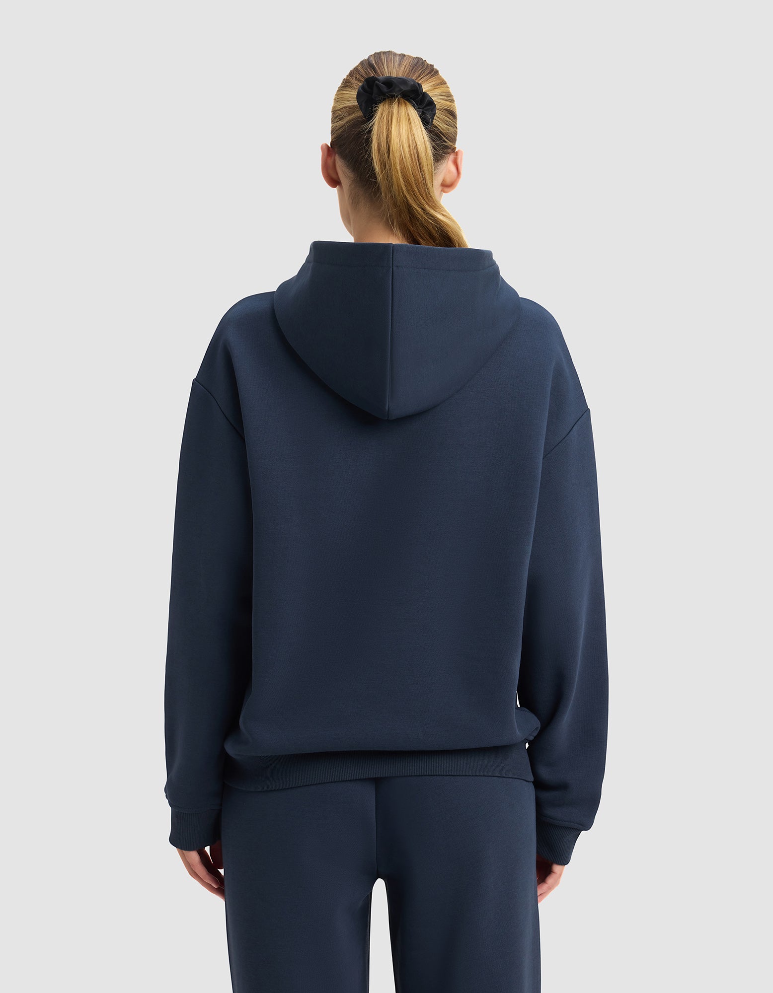 A woman with blonde hair in a ponytail wears the Cozy Earth Women's Cityscape 2.0 Hoodie and matching pants, facing away from the camera against a plain light gray background.|Color:Eclipse