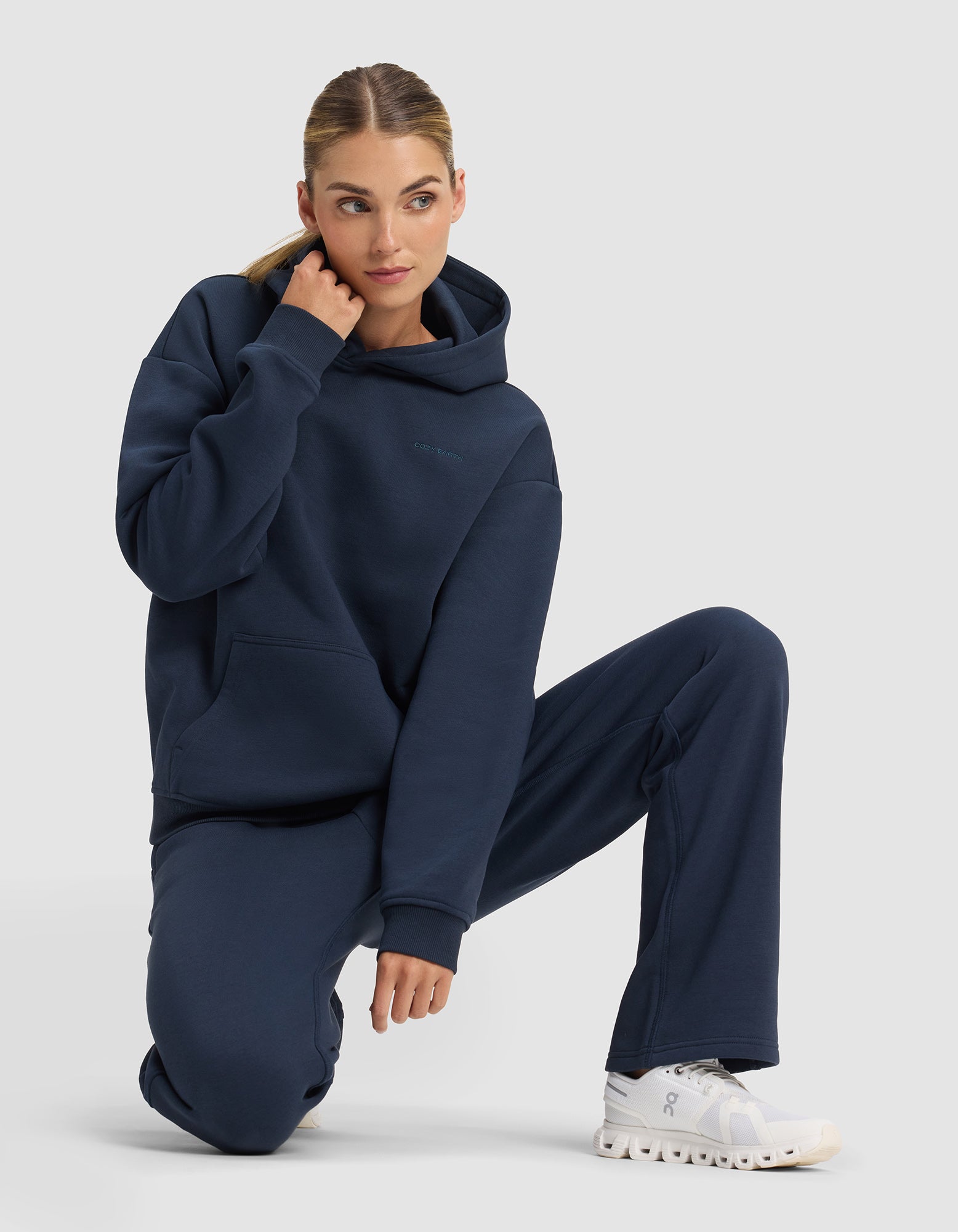 A woman with blonde hair tied back kneels on one knee against a plain light gray background, wearing the Cozy Earth Women's Cityscape 2.0 Hoodie in navy blue with matching sweatpants and white athletic shoes.|Color:Eclipse