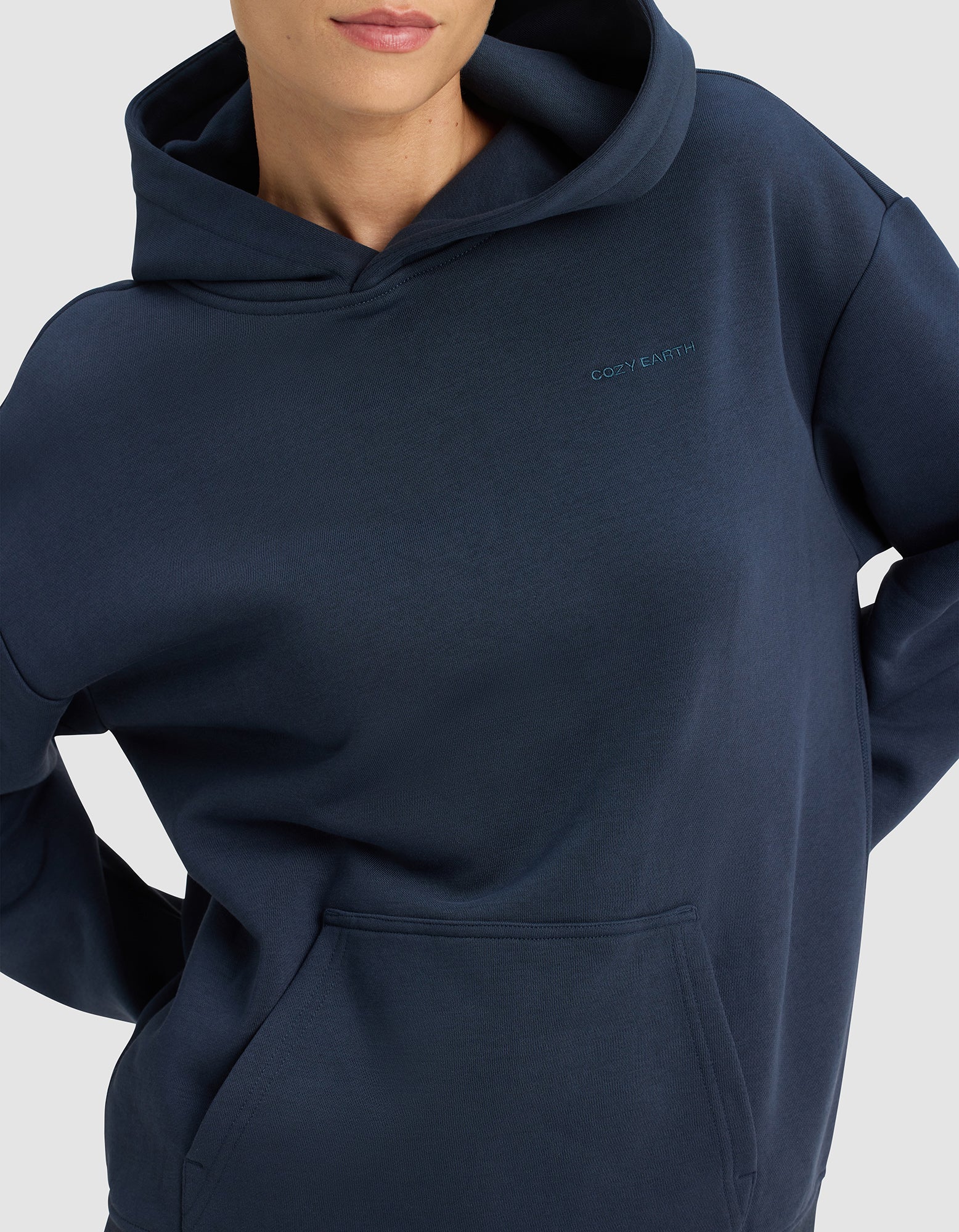 A woman wears the Cozy Earth Women’s Cityscape 2.0 Hoodie in dark blue, featuring "COZY EARTH" embroidered on the chest, standing with hands on hips against a plain light background.|Color:Eclipse