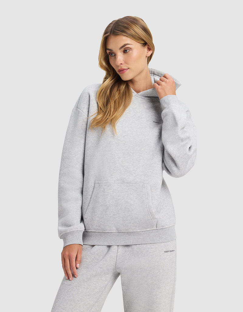 A woman wears the Cozy Earth Women's Cityscape 2.0 Hoodie in light gray, standing against a plain background and looking to the side as she adjusts her collar.