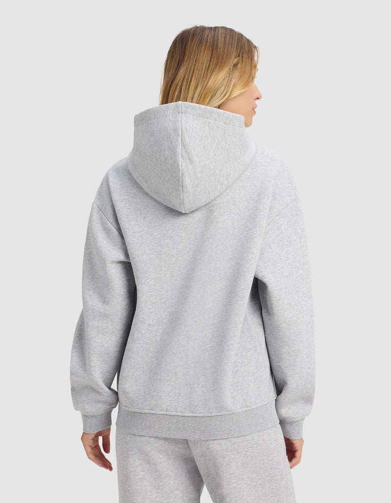 A woman with shoulder-length blonde hair is seen from behind wearing the Cozy Earth Women's Cityscape 2.0 Hoodie in light gray, with matching sweatpants, against a plain white background.