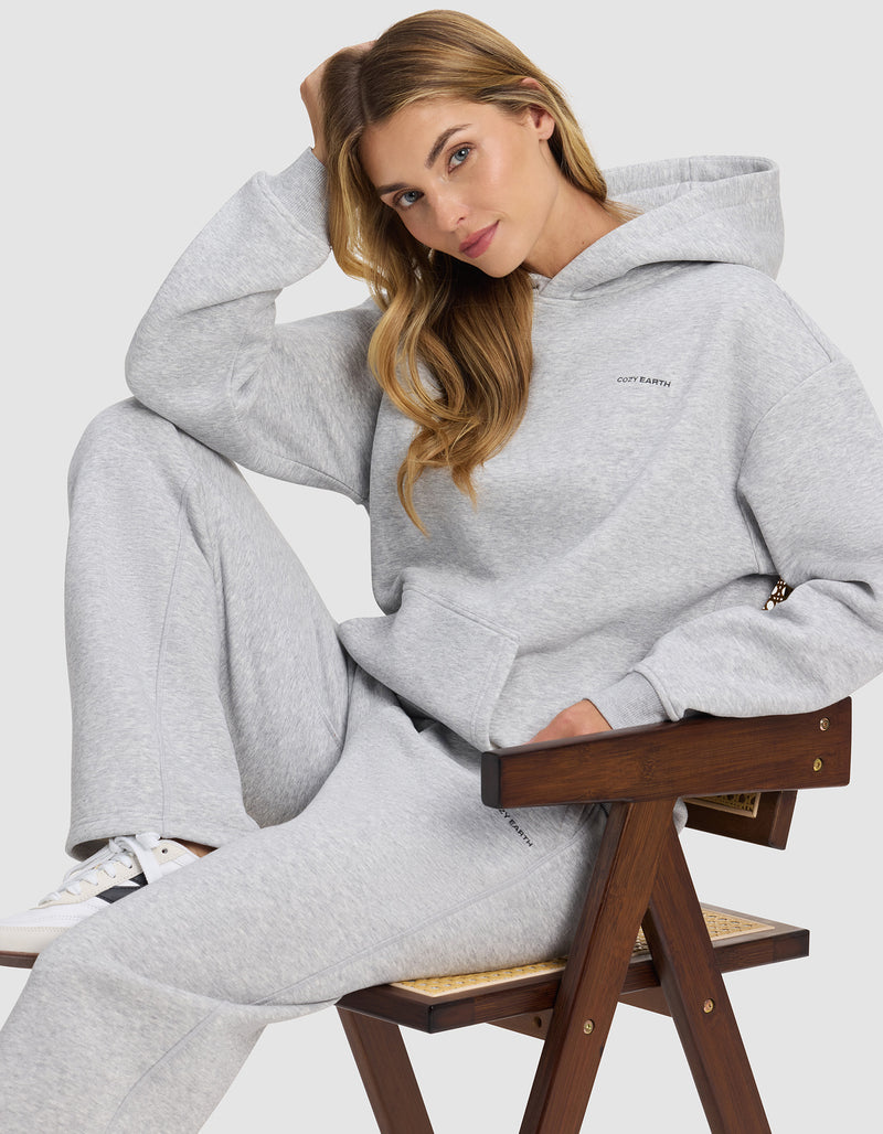 A woman with long blonde hair sits on a wooden chair, wearing the Cozy Earth Women's Cityscape 2.0 Hoodie in light gray, with matching sweatpants. She rests one arm on her head and gazes relaxingly at the camera.