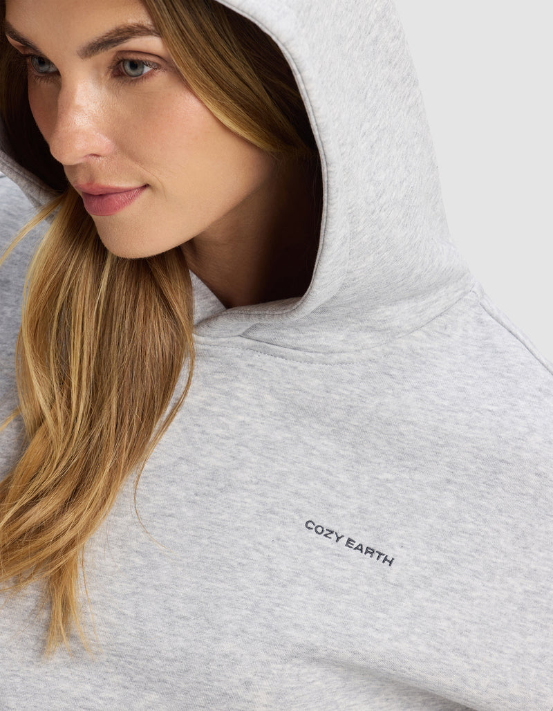 A woman with long blonde hair wears the Cozy Earth Women's Cityscape 2.0 Hoodie in light gray, featuring a small black "COZY EARTH" logo on the chest. She looks slightly to the side with a gentle smile.