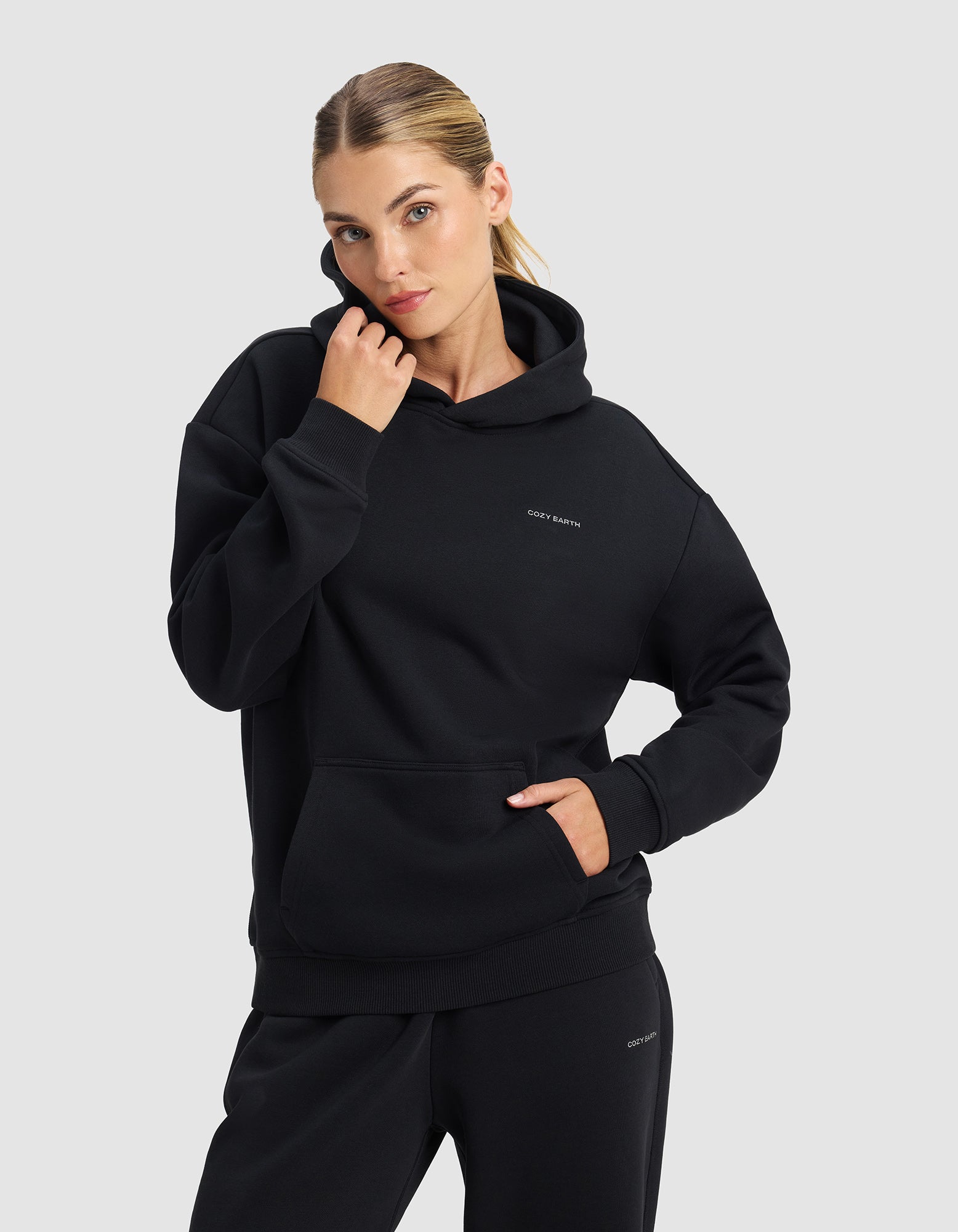 A woman with blonde hair in a ponytail models the Cozy Earth Women's Cityscape 2.0 Hoodie in Jet Black, posing with one hand near her face and the other in her pocket against a plain light grey background.|Color:Jet Black