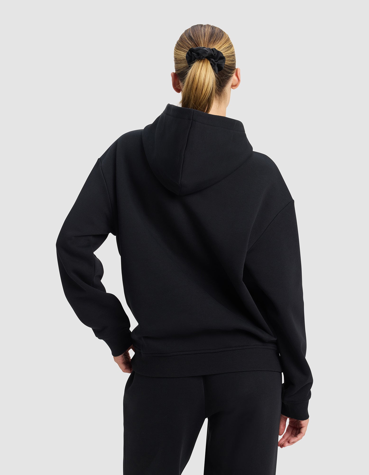 A woman with blonde hair in a ponytail, tied with a black scrunchie, wears the Cozy Earth Women's Cityscape 2.0 Hoodie and black pants. She faces away from the camera against a plain light gray background.