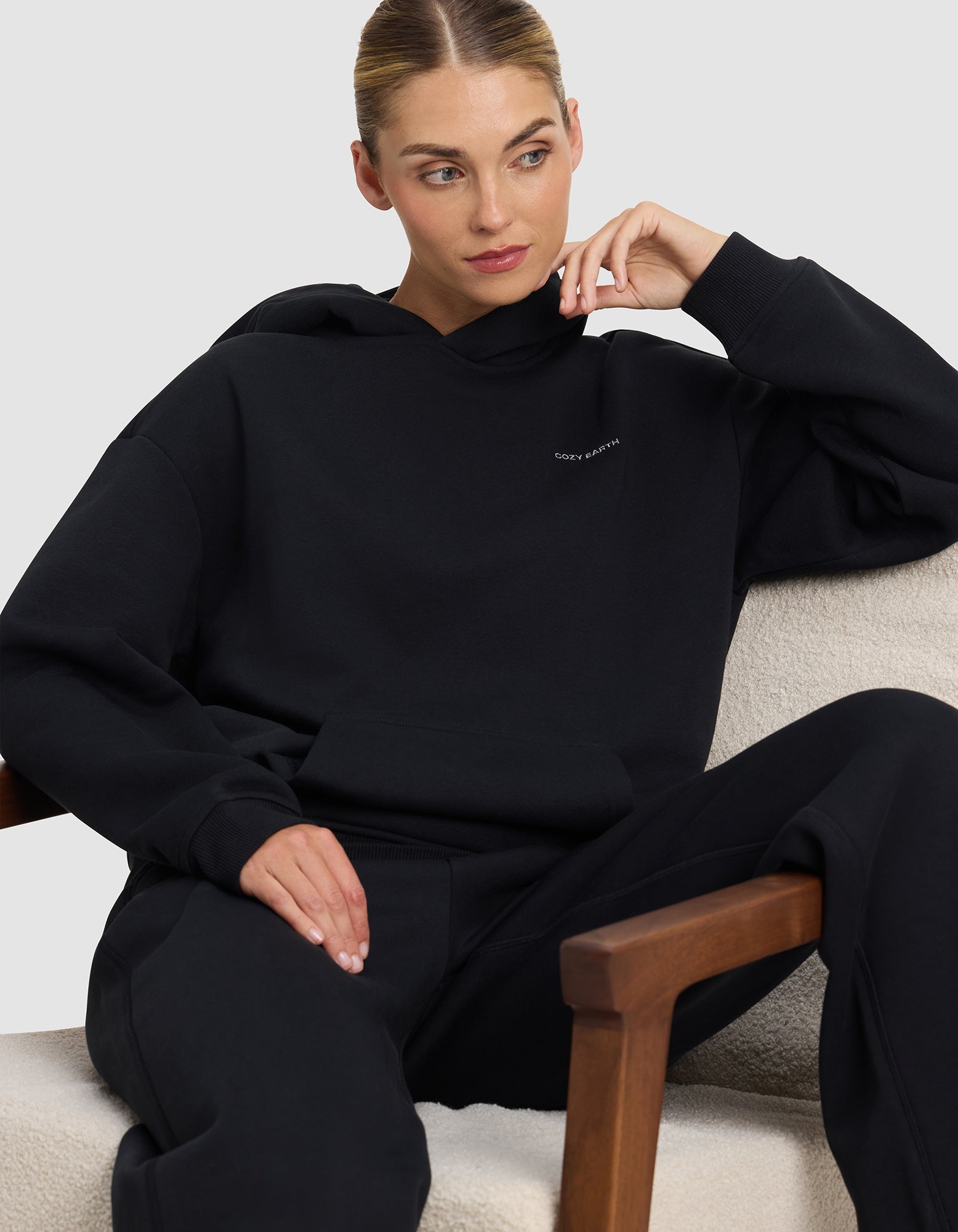 A woman with her hair tied back sits on a beige chair, wearing the Cozy Earth Women's Cityscape 2.0 Hoodie in black and matching pants. She rests her elbow on the chair’s arm, gazing to the side with a relaxed expression.
