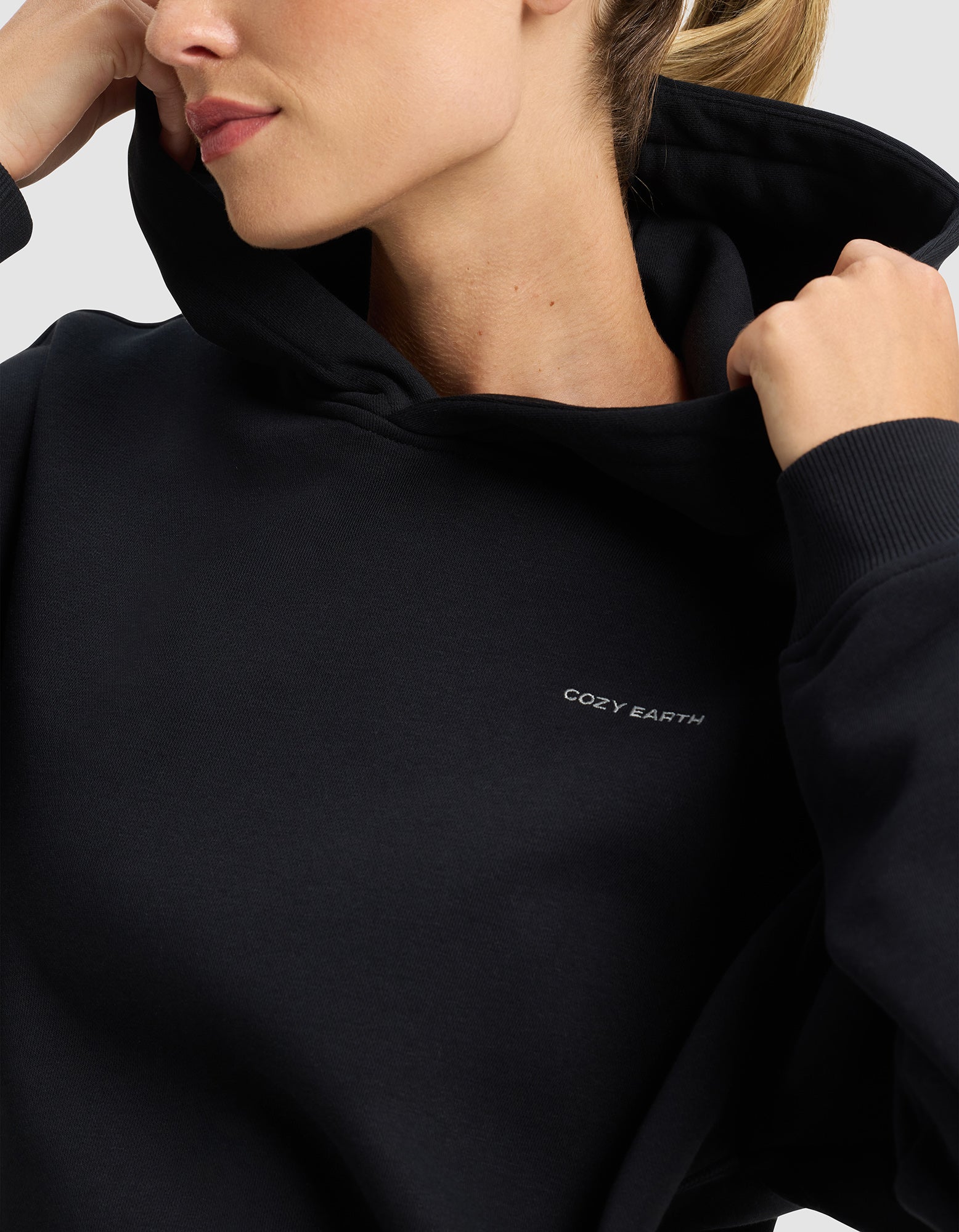 A person wears the Women's Cityscape 2.0 Hoodie by Cozy Earth, hood partially up and holding the collar. The black hoodie features "COZY EARTH" in small white letters on the chest, set against a plain light background.