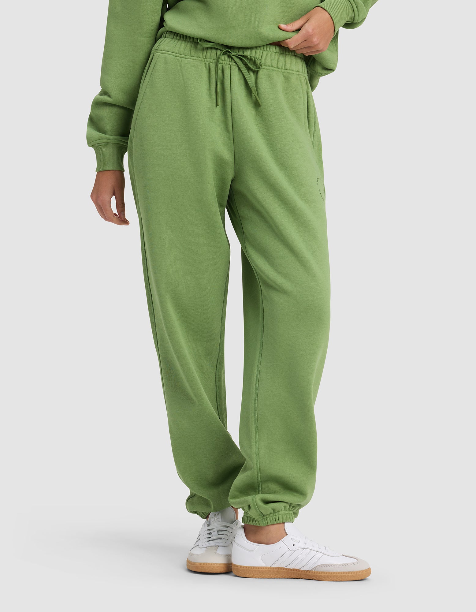A person wears Cozy Earth's Women's Cityscape 2.0 Sweatpant—a green pair with an elastic waistband and drawstring—styled with a matching sweatshirt and white sneakers, standing against a plain light background.|Color:Clover