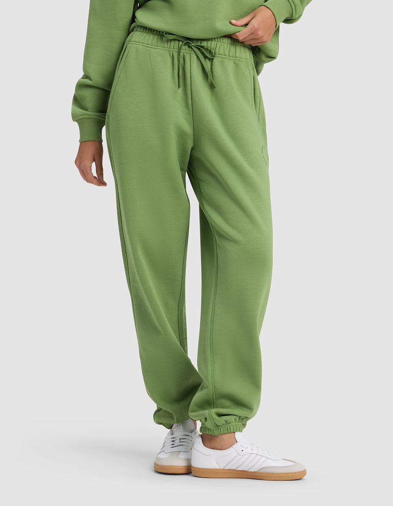 A person wears Cozy Earth's Women's Cityscape 2.0 Sweatpant—a green pair with an elastic waistband and drawstring—styled with a matching sweatshirt and white sneakers, standing against a plain light background.