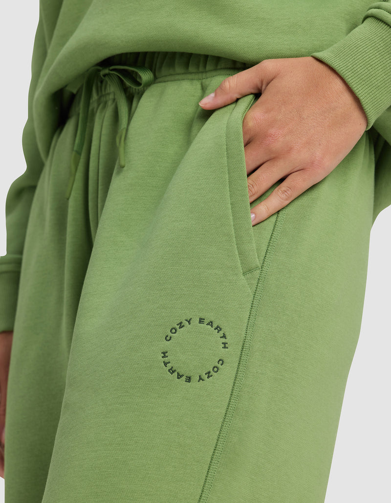 A person wears Cozy Earth's Women's Cityscape 2.0 Sweatpant in light green with a matching sweatshirt, one hand in their pocket. "COZY EARTH" is printed in a circle near the sweatpant’s pocket.