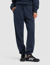 A woman wearing Cozy Earth Women's Cityscape 2.0 Sweatpant stands with one hand in her pocket against a plain light background, paired with black sneakers featuring white stripes.|Color:Eclipse