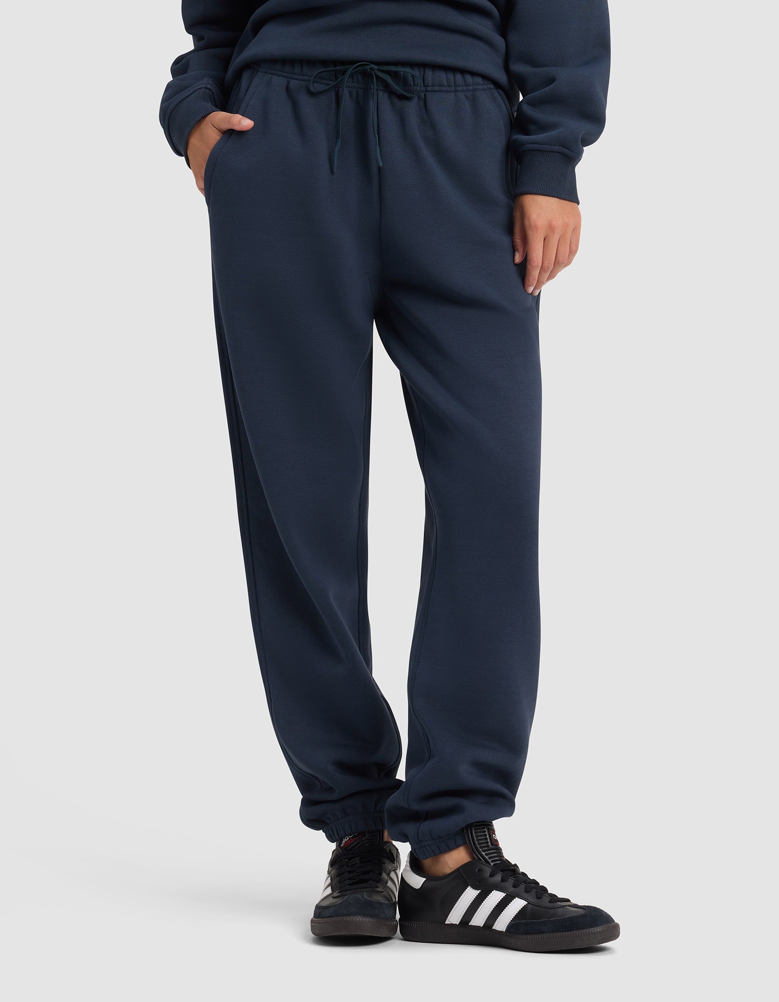 A woman wearing Cozy Earth Women's Cityscape 2.0 Sweatpant stands with one hand in her pocket against a plain light background, paired with black sneakers featuring white stripes.|Color:Eclipse