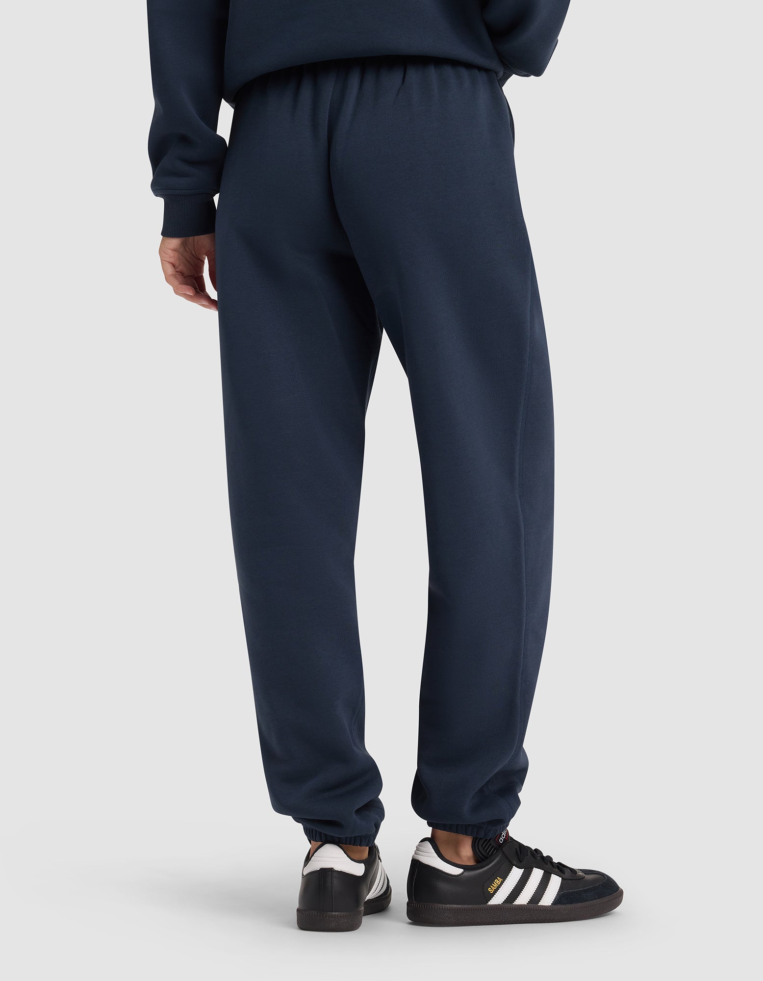 A person is seen from behind wearing Cozy Earth Women's Cityscape 2.0 Sweatpants in dark blue with a matching sweatshirt, paired with black Adidas sneakers featuring white stripes and gold branding.