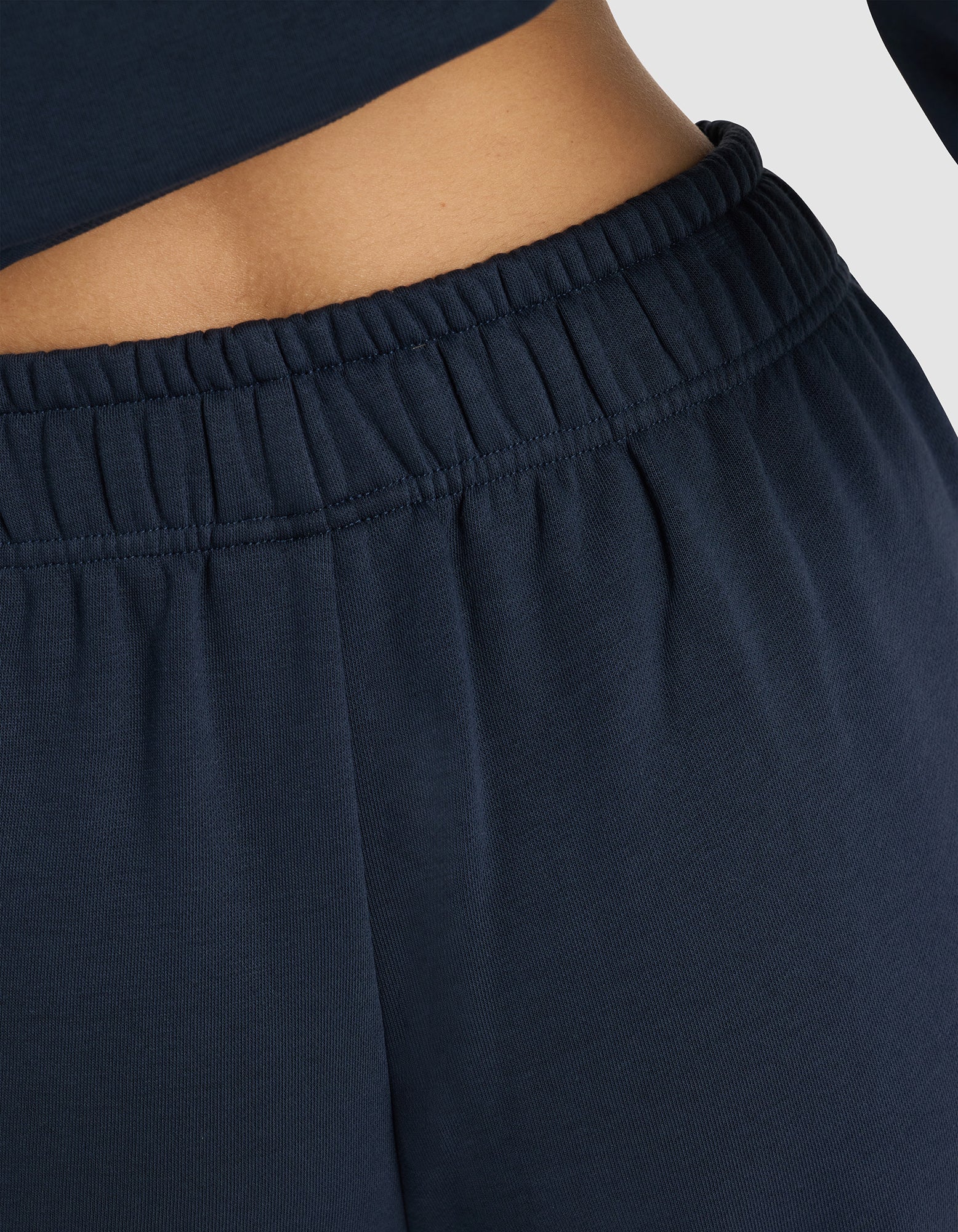 Close-up of a person wearing Cozy Earth Women's Cityscape 2.0 Sweatpant in navy blue with a high-waisted elastic waistband and matching top, highlighting the waistband and lower torso against a light background.|Color:Eclipse
