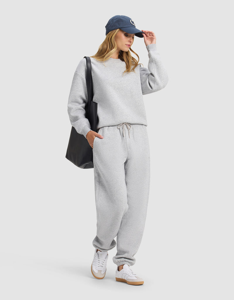 A woman in Cozy Earth's Cityscape 2.0 Sweatpant, a gray sweatshirt, white sneakers, navy blue cap, and carrying a large black tote poses against a plain background with one hand in her pocket and the other adjusting her cap.