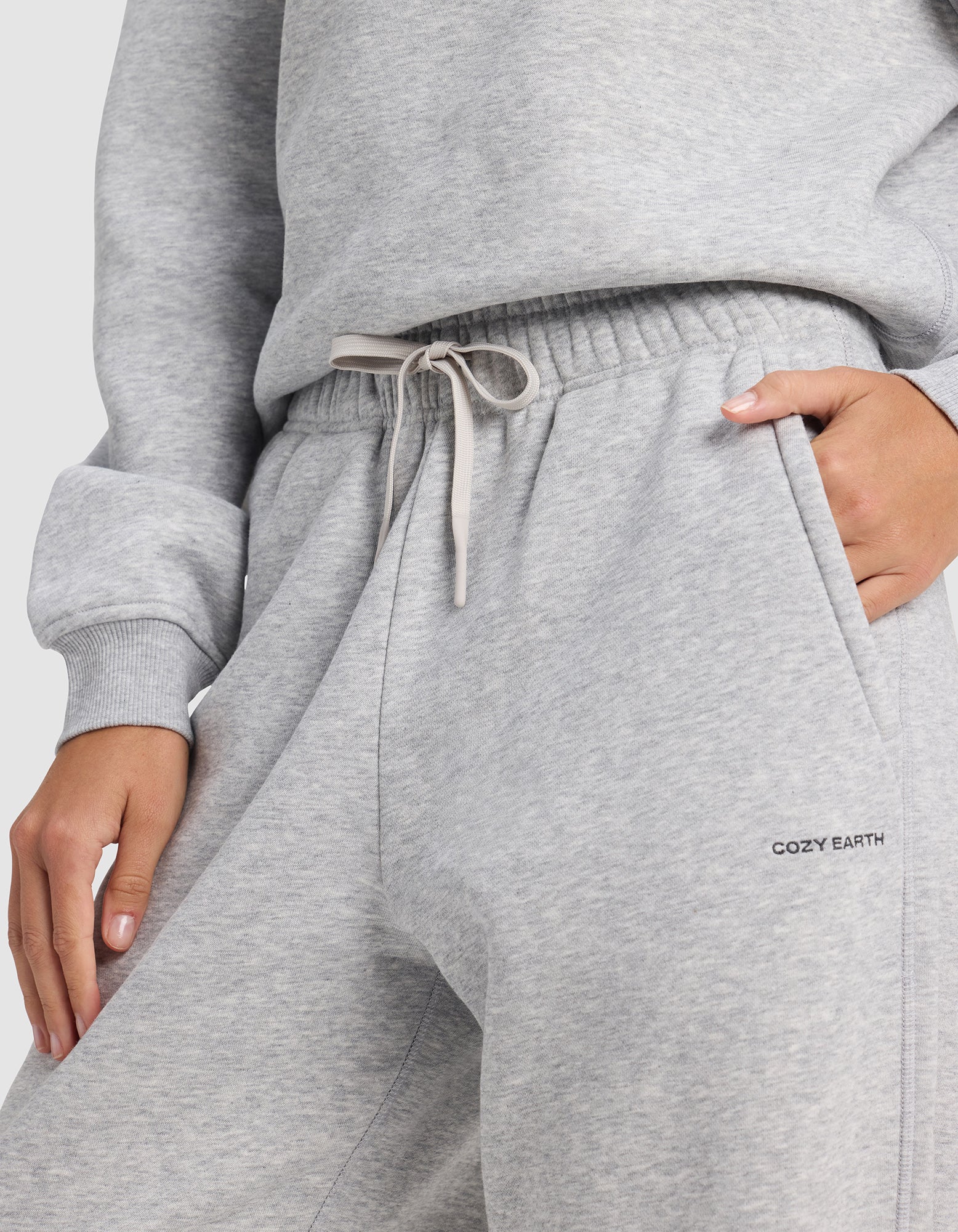 A person wears light gray Women's Cityscape 2.0 Sweatpants by Cozy Earth, featuring an elastic waistband with drawstring, one hand in a pocket, and "COZY EARTH" printed on the side.